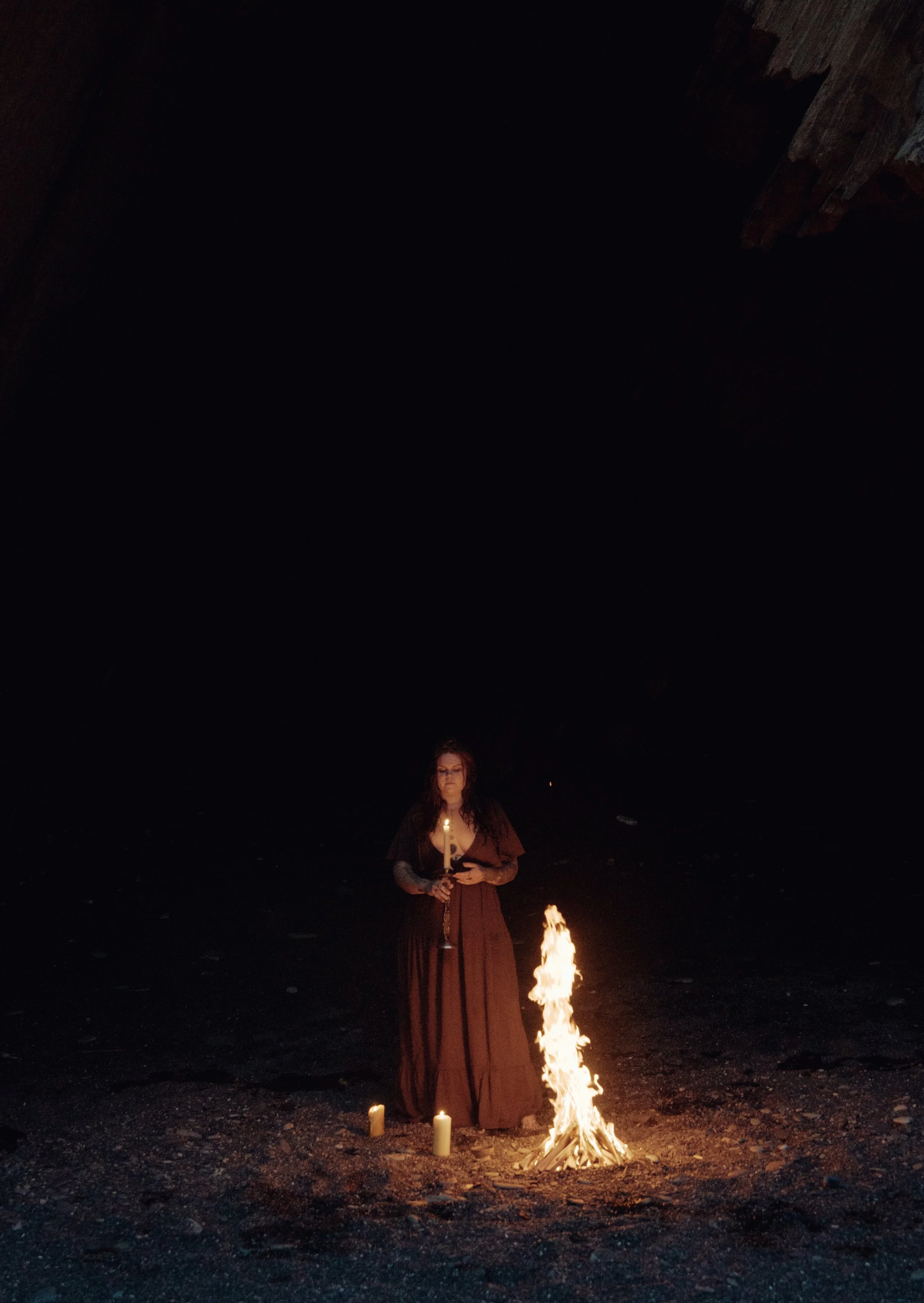 A woman wearing a long dress stands in a dark, cavernous space, holding a lit candle in each hand. In front of her is a small fire on the ground, with a few candles placed nearby.