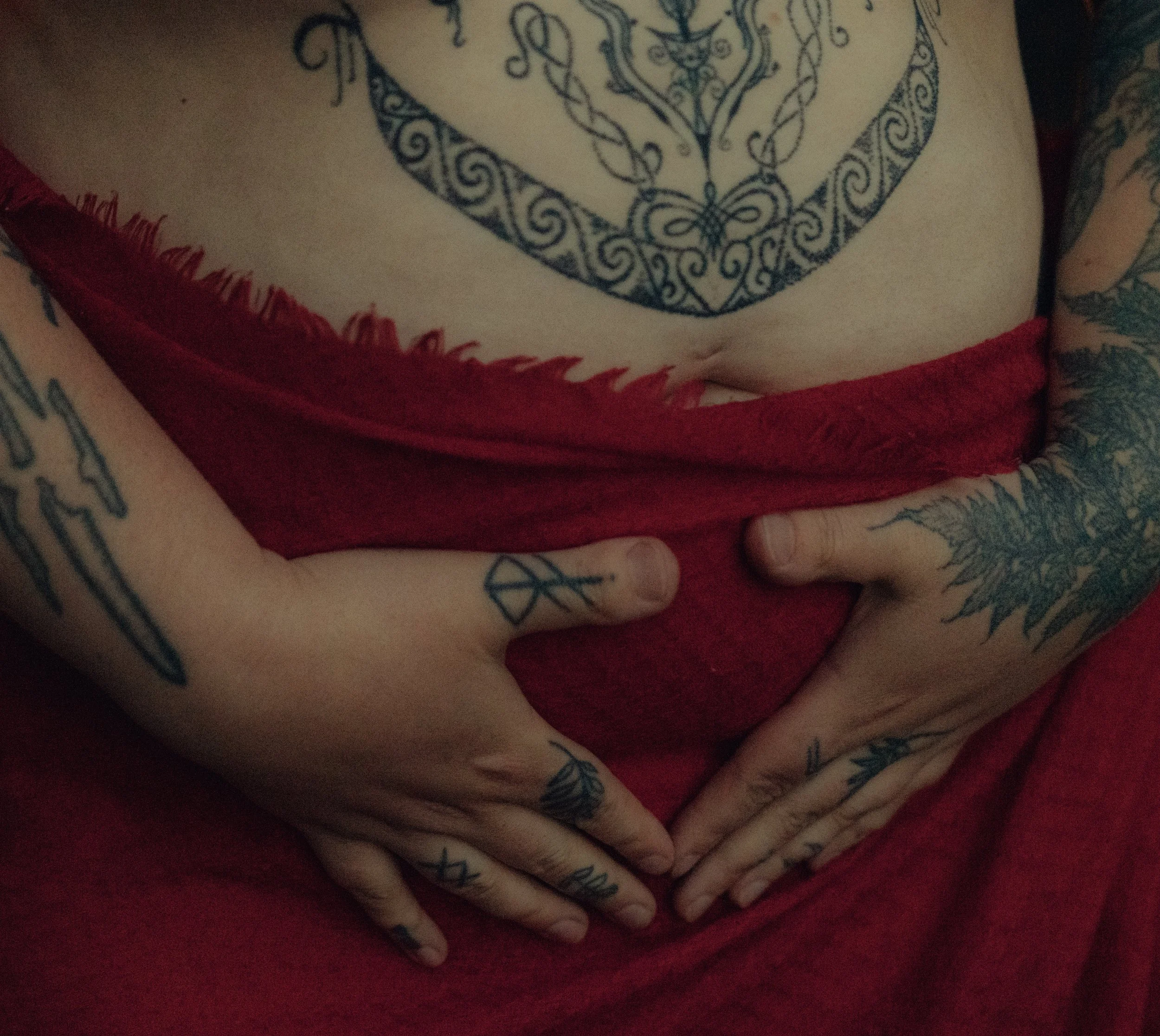 Close-up of a person's tattooed hands resting on their stomach, with a large tattoo on their chest visible. The person is wearing a red shirt.
