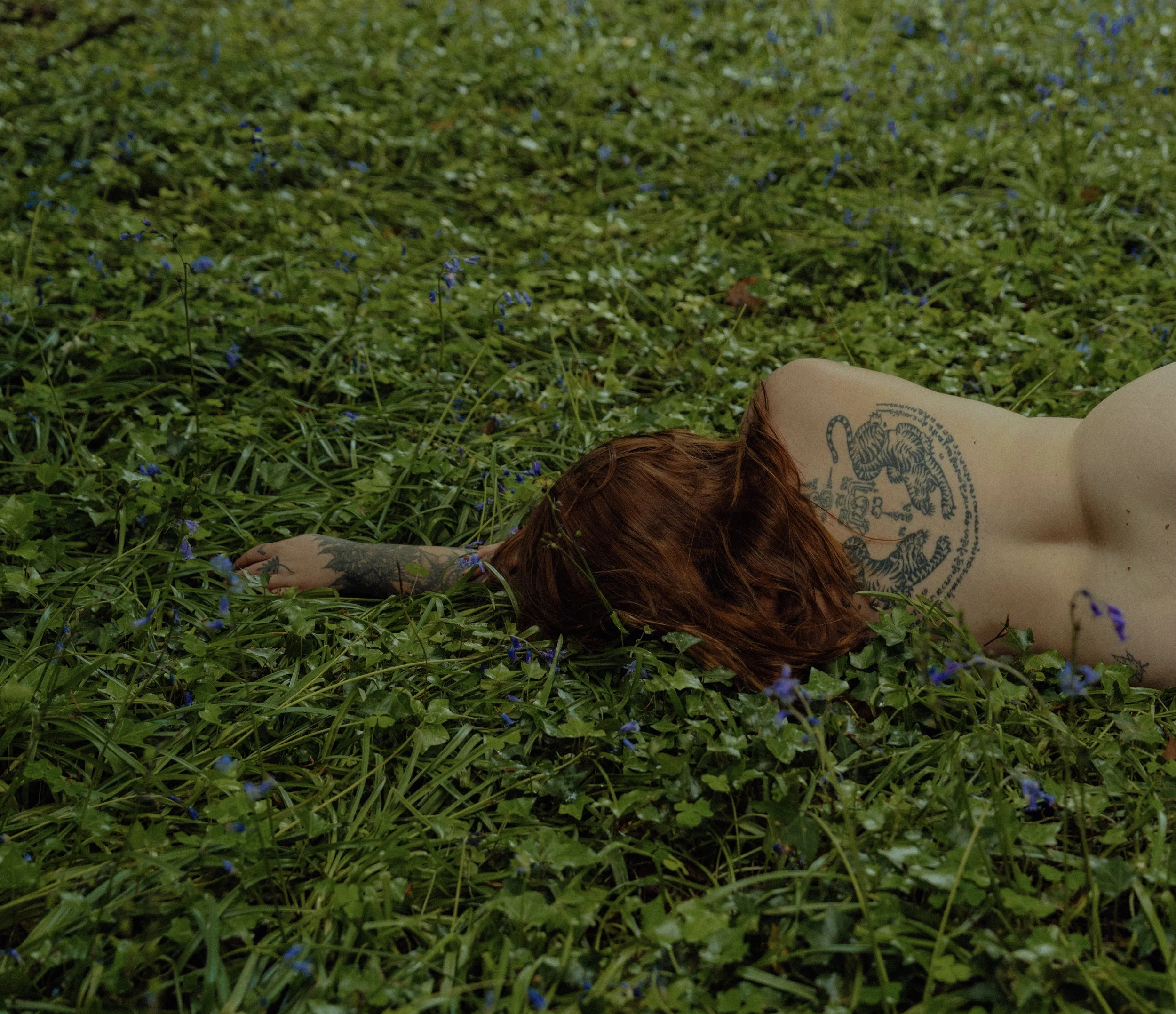 Person with red hair and tattoos lying face down in a field of green grass and blue flowers.