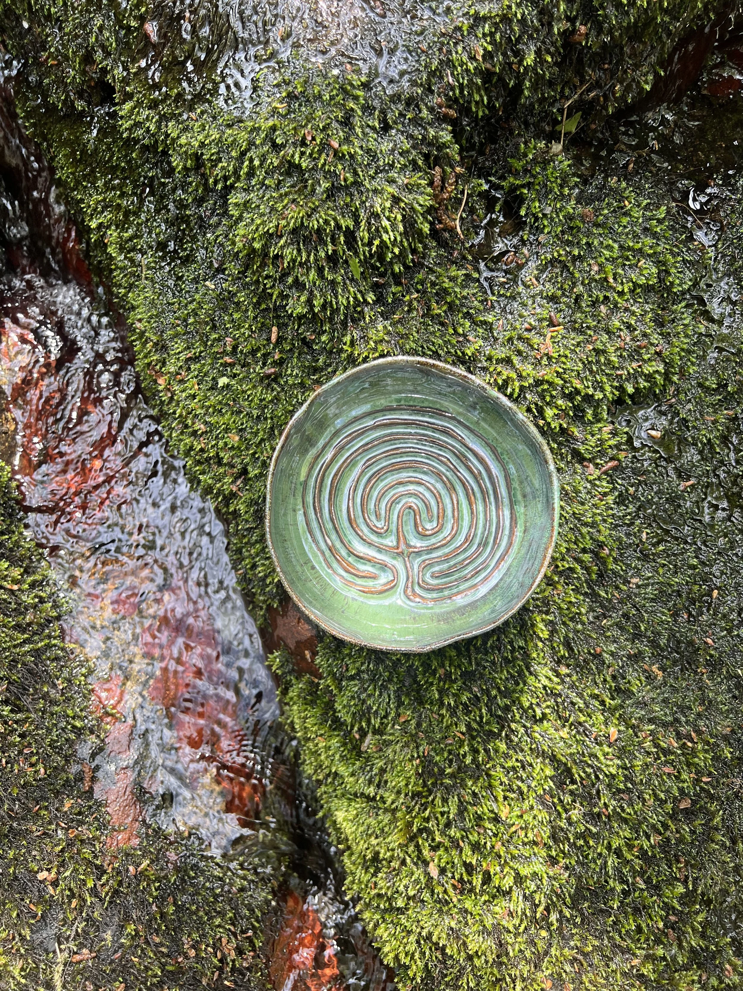 Close-up of a moss-covered rock with an embedded circular glass piece featuring a labyrinth pattern, and a small stream flowing beside it.