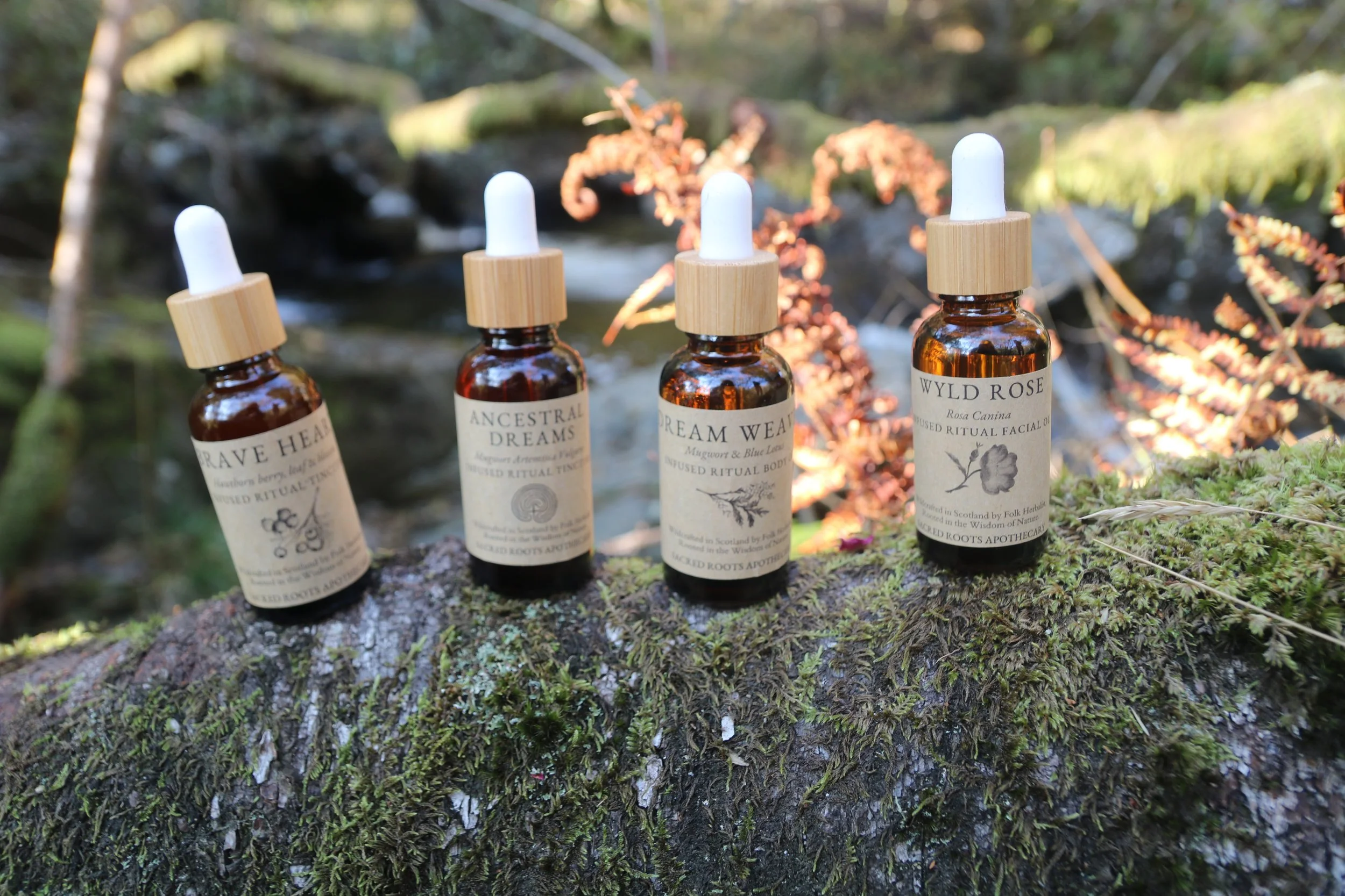 Four small amber glass bottles with white dropper caps lined up on a moss-covered log in a forest setting, each labeled with different herbal or botanical names.