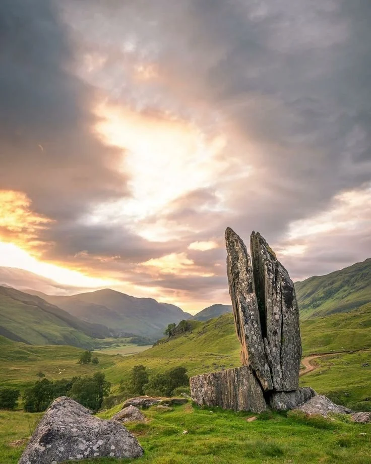 A large, vertical standing stone sculpture resembling an abstract pair of fingers, set in a lush green valley with rolling hills and mountains in the background during sunset.