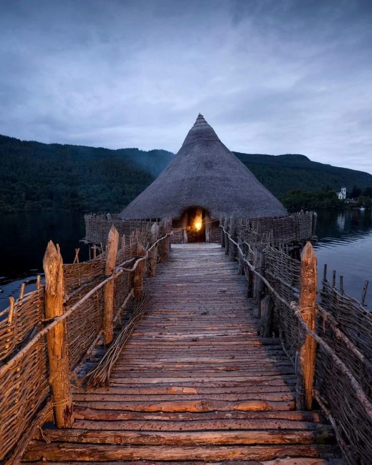 A wooden bridge leading to a thatched-roof hut by a lake, with mountains in the background and a glowing light inside the hut.