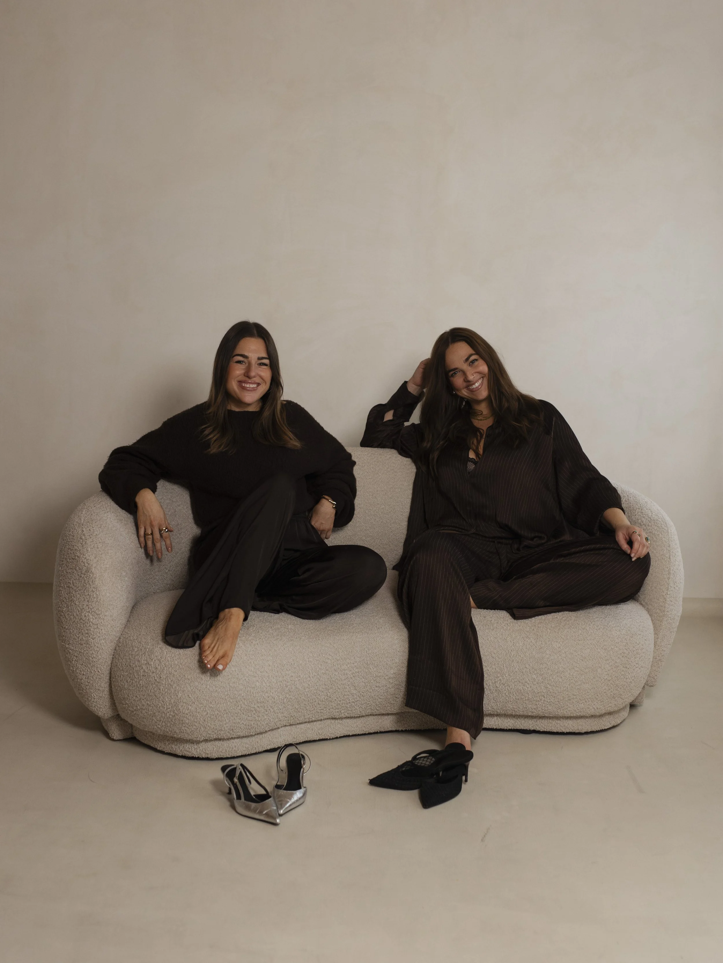 Zwei lachende Frauen sitzen entspannt auf einem weißen, gemütlichen Sofa gegenüber einer weißen Wand. Darunter liegen zwei Paar High Heels, eines in Schwarz und das andere in Silber.