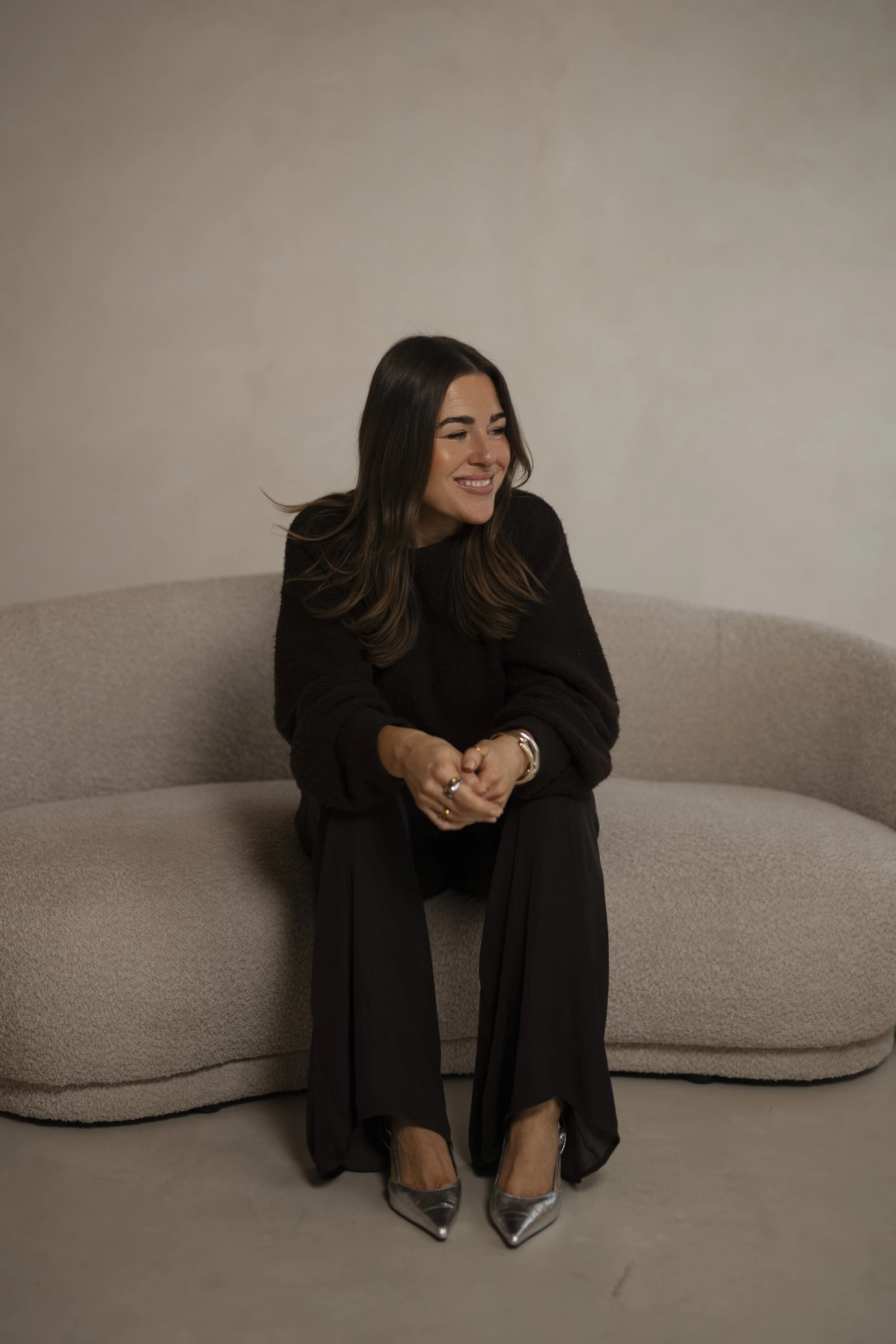 Eine Frau sitzt auf einem beige Couch, trägt ein schwarzes Oberteil und schwarze Hosen, sowie silberne High Heels, lächelnd und schaut zur Seite.