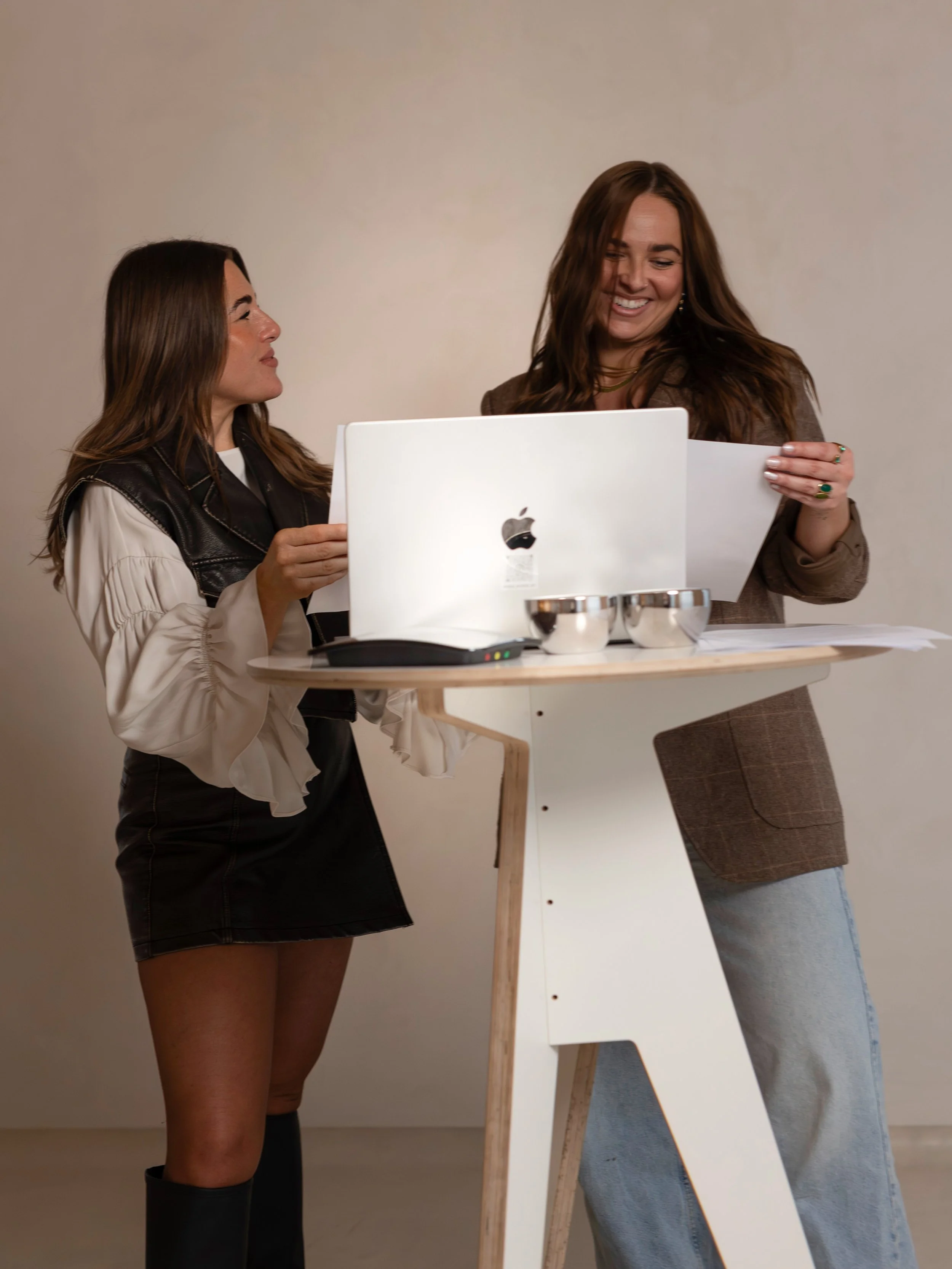 Zwei Frauen lachen und diskutieren bei einer Arbeitssitzung, umgeben von einem Laptop, Dokumenten und Schalen auf einem Stehpult vor neutralem Hintergrund.