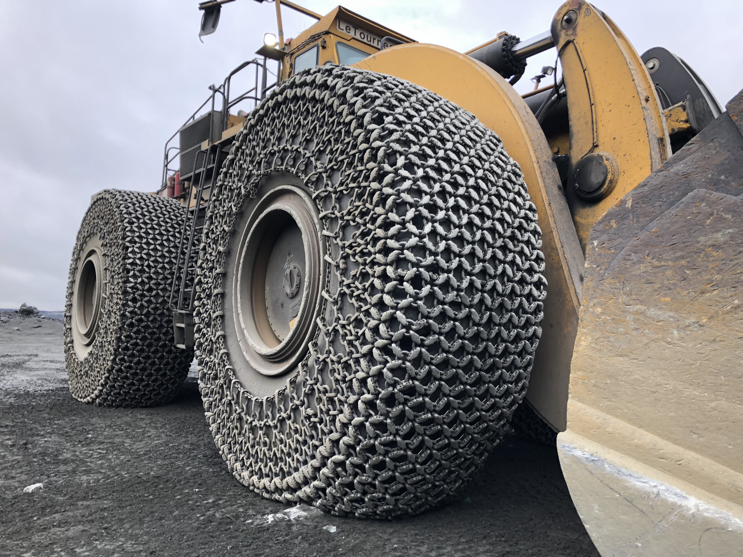 Close-up of large construction tires with heavy-duty metal chains wrapped around the tires to provide traction on rough terrain, with a yellow construction vehicle and a cloudy sky in the background.