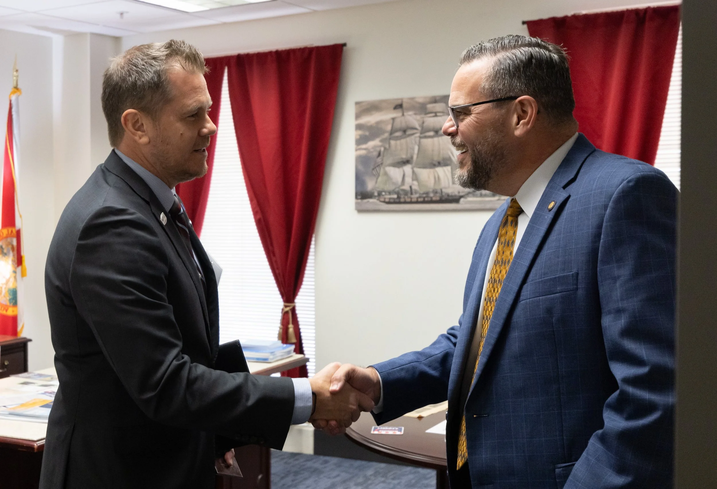 Two men in suits shaking hands in an office with red curtains, a picture of a sailing ship, and a flag in the background.
