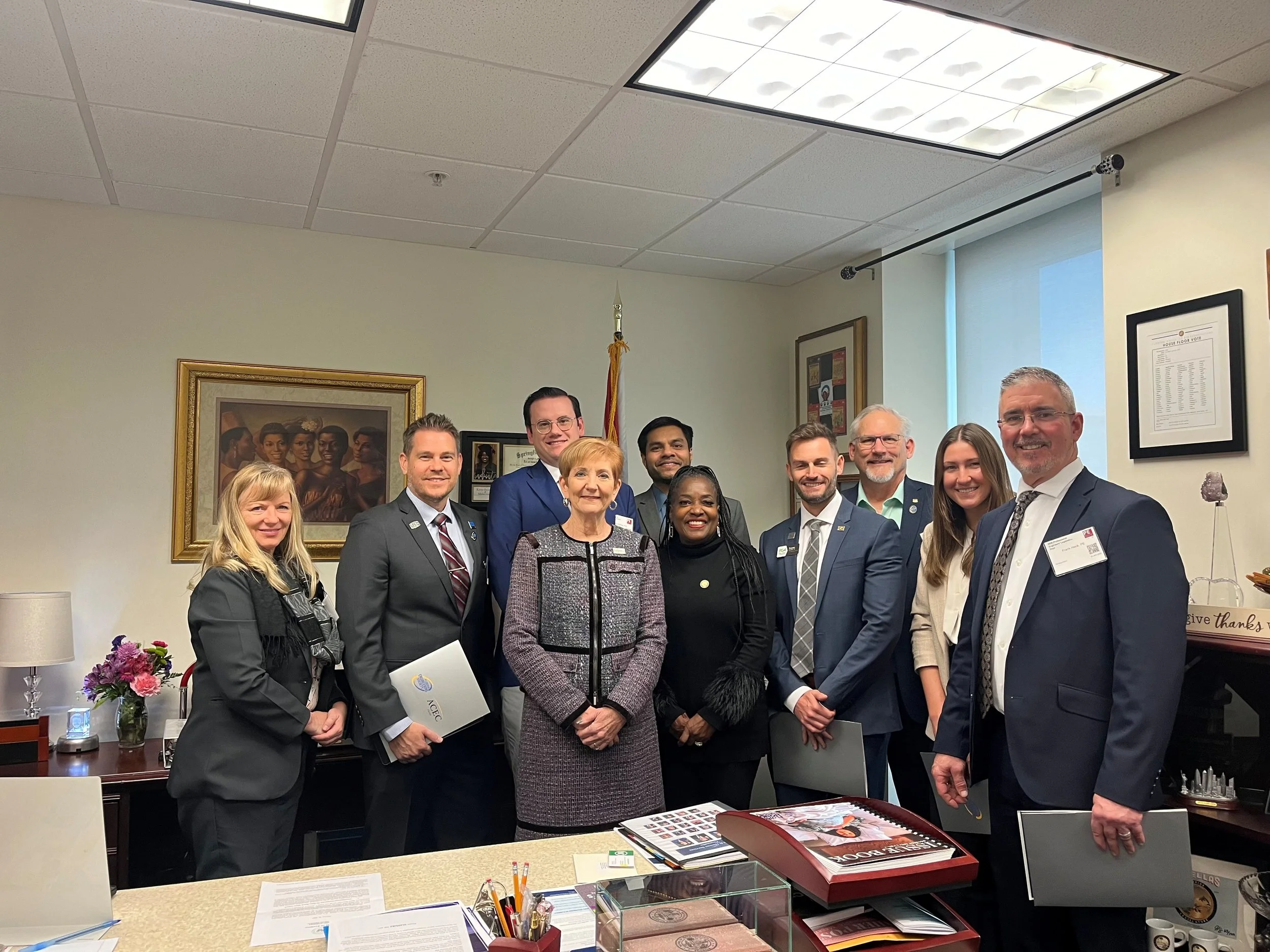 A group of eleven professionally dressed individuals posing in an office, with American flags, framed pictures and certificates on the wall, and a desk with documents and flowers in the foreground.