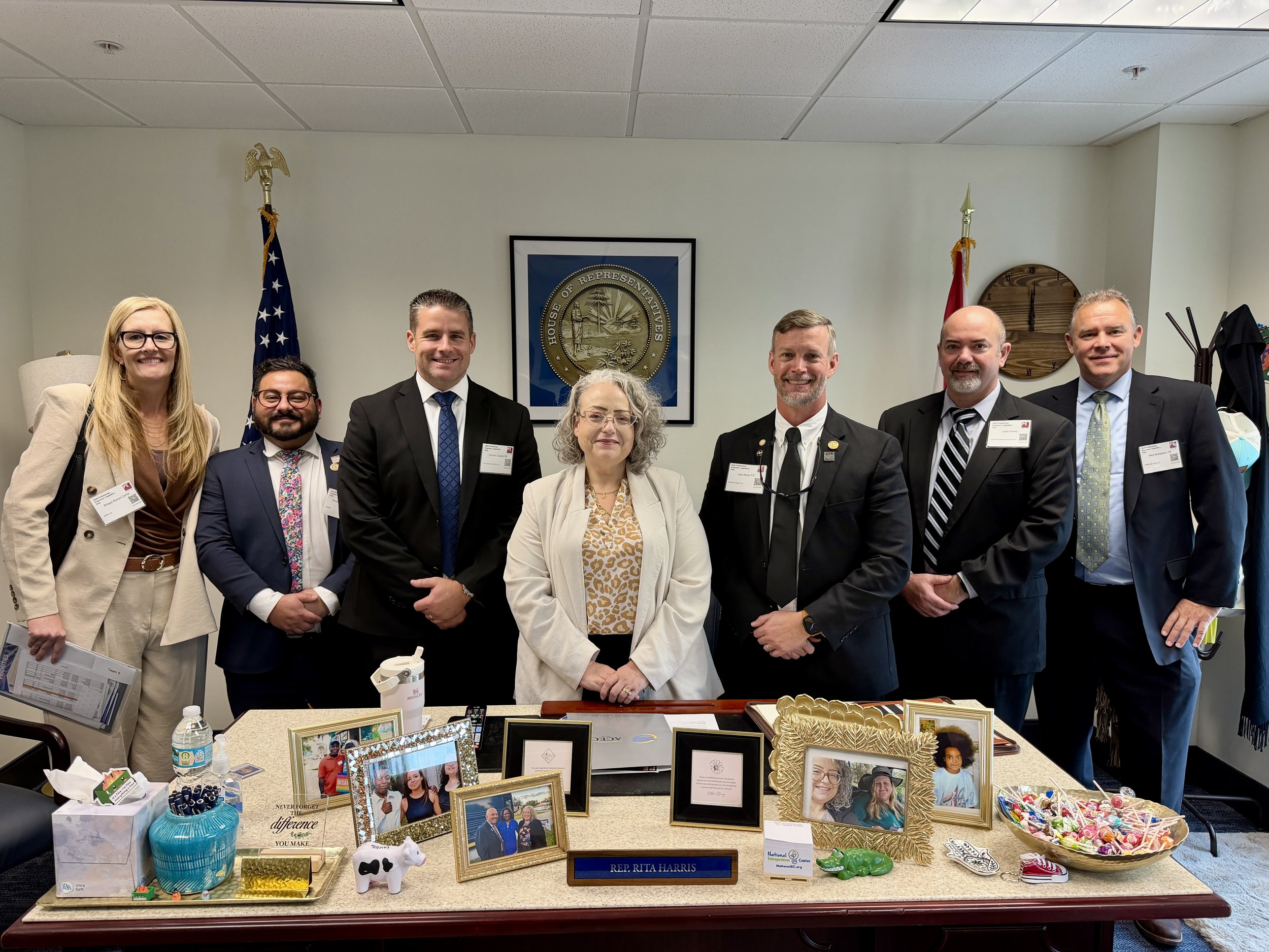 A group of nine professionally dressed people standing behind a table decorated with framed photos, a bowl of candy, and various decorative items, all in an office with a framed seal and flags on the wall.