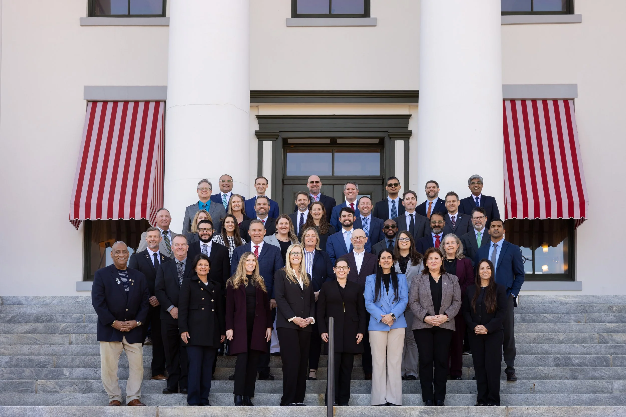 Group of professionally dressed people standing on steps in front of a building with large columns and red-and-white striped awnings.