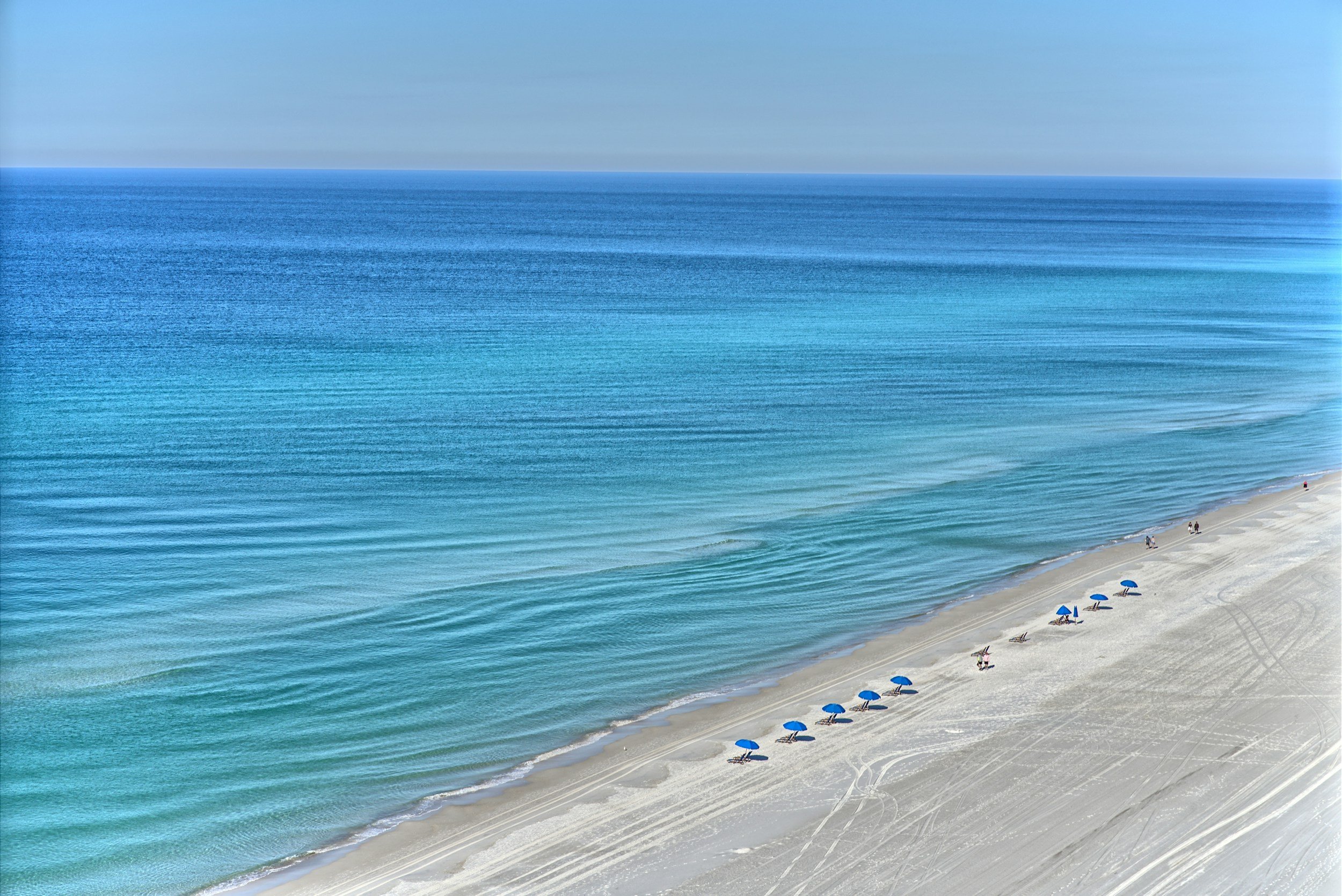 A sandy beach with blue umbrellas and a few people walking along the shoreline, with calm blue ocean water and a clear sky in the background.