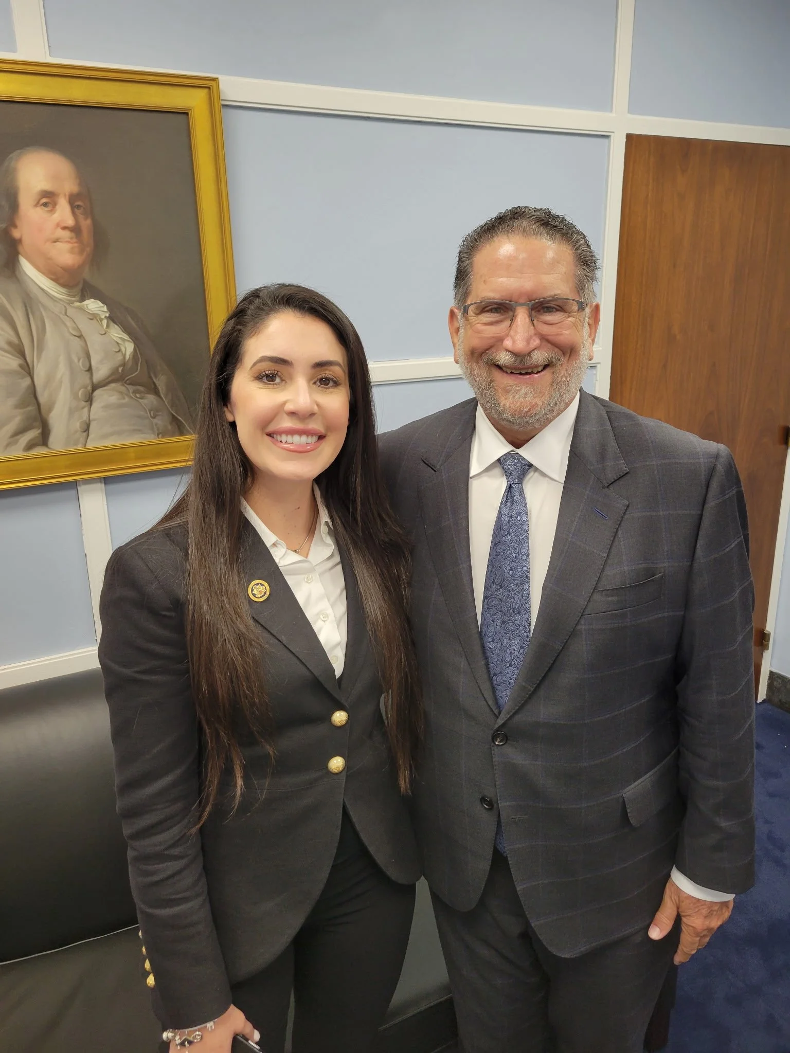 A young woman and an older man in suits are standing and smiling in a room with blue walls, a black leather couch, and a framed portrait of a man in historical clothing hanging on the wall behind them.