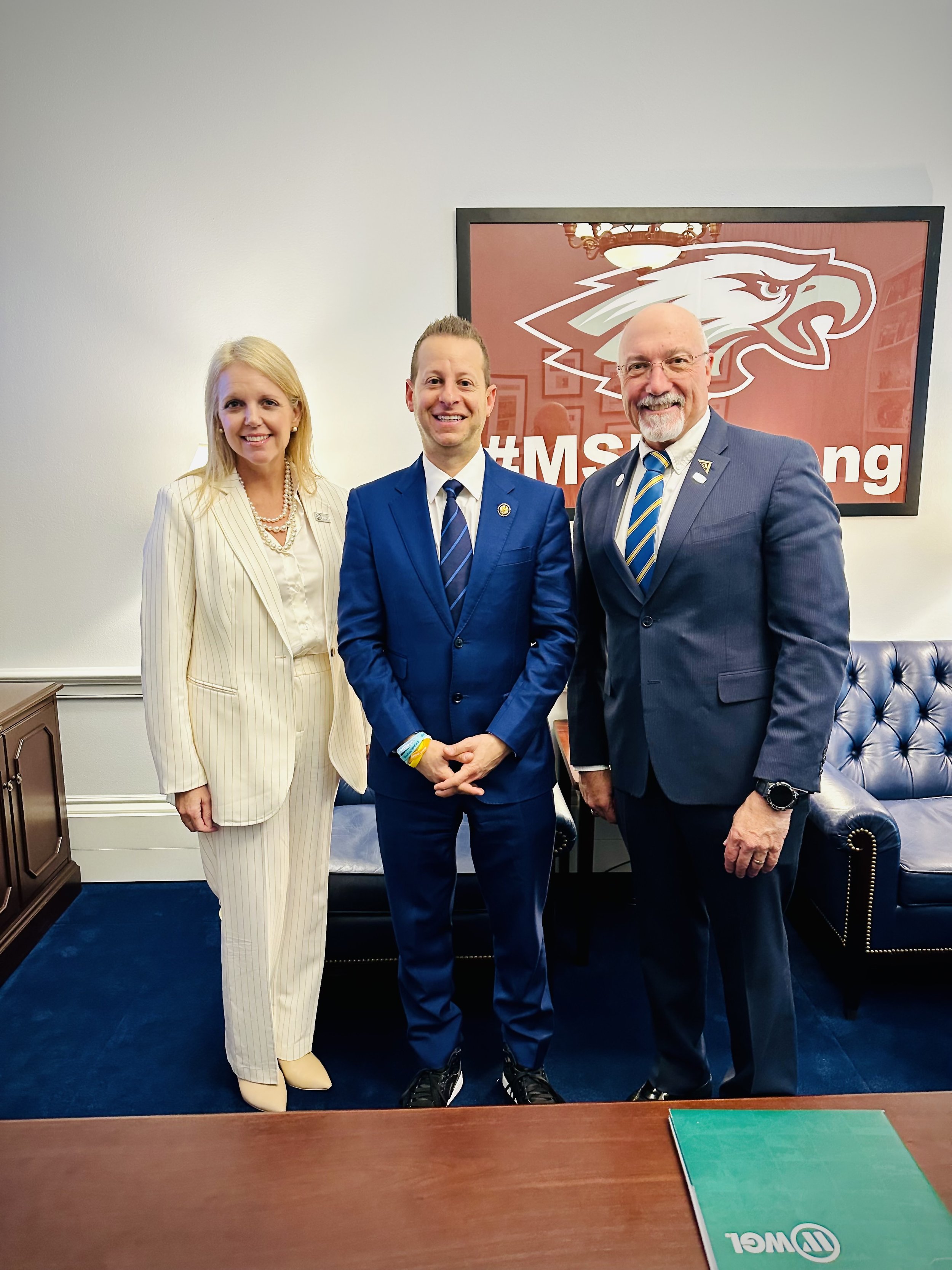 Three people in business suits standing in an office, smiling for a photo, with a large framed logo of a stylized eagle behind them and a hashtag #MSI in the background.