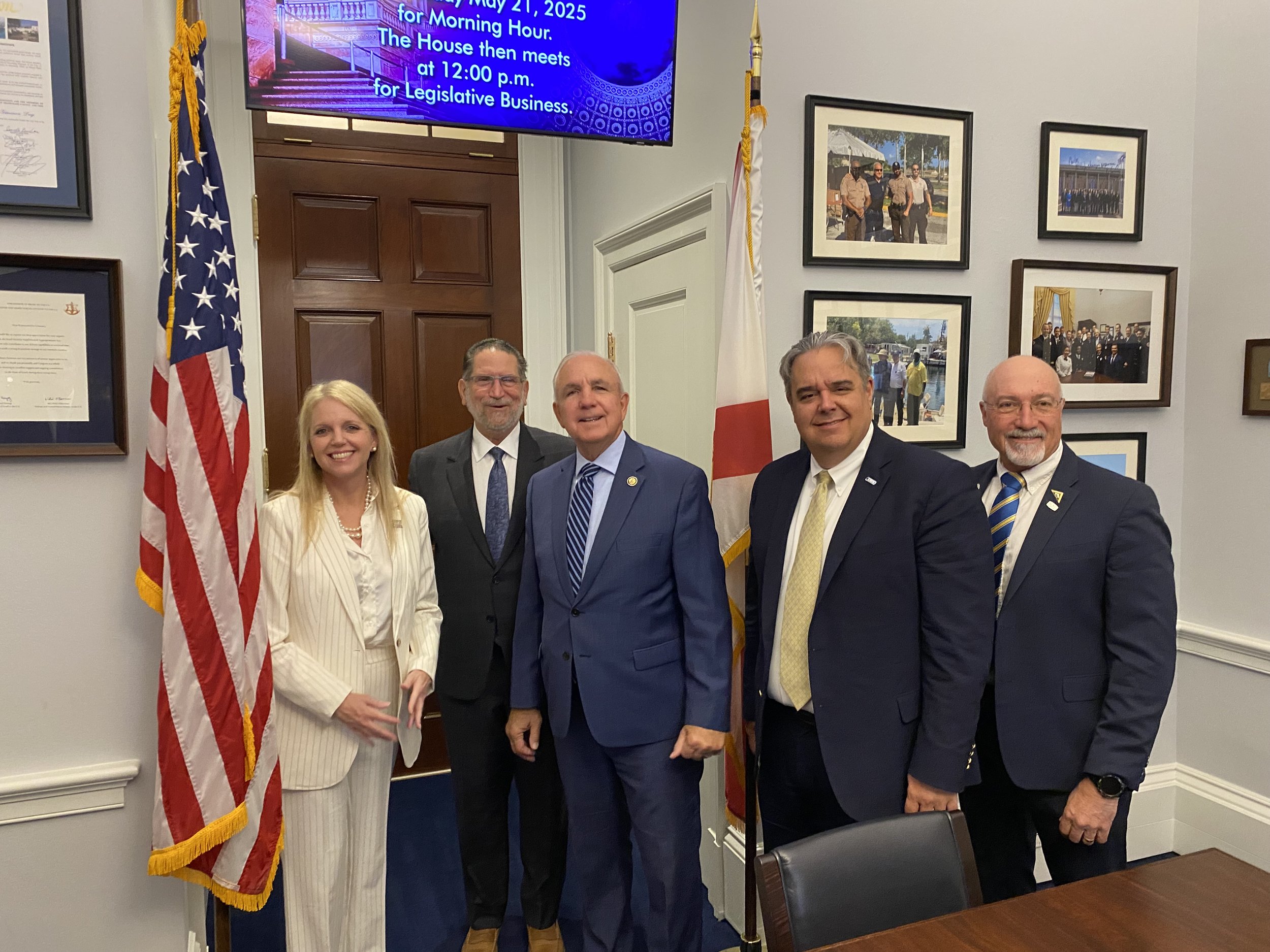 Six people standing in a room with American and state flags, smiling for a photo, with framed pictures on the wall behind them.