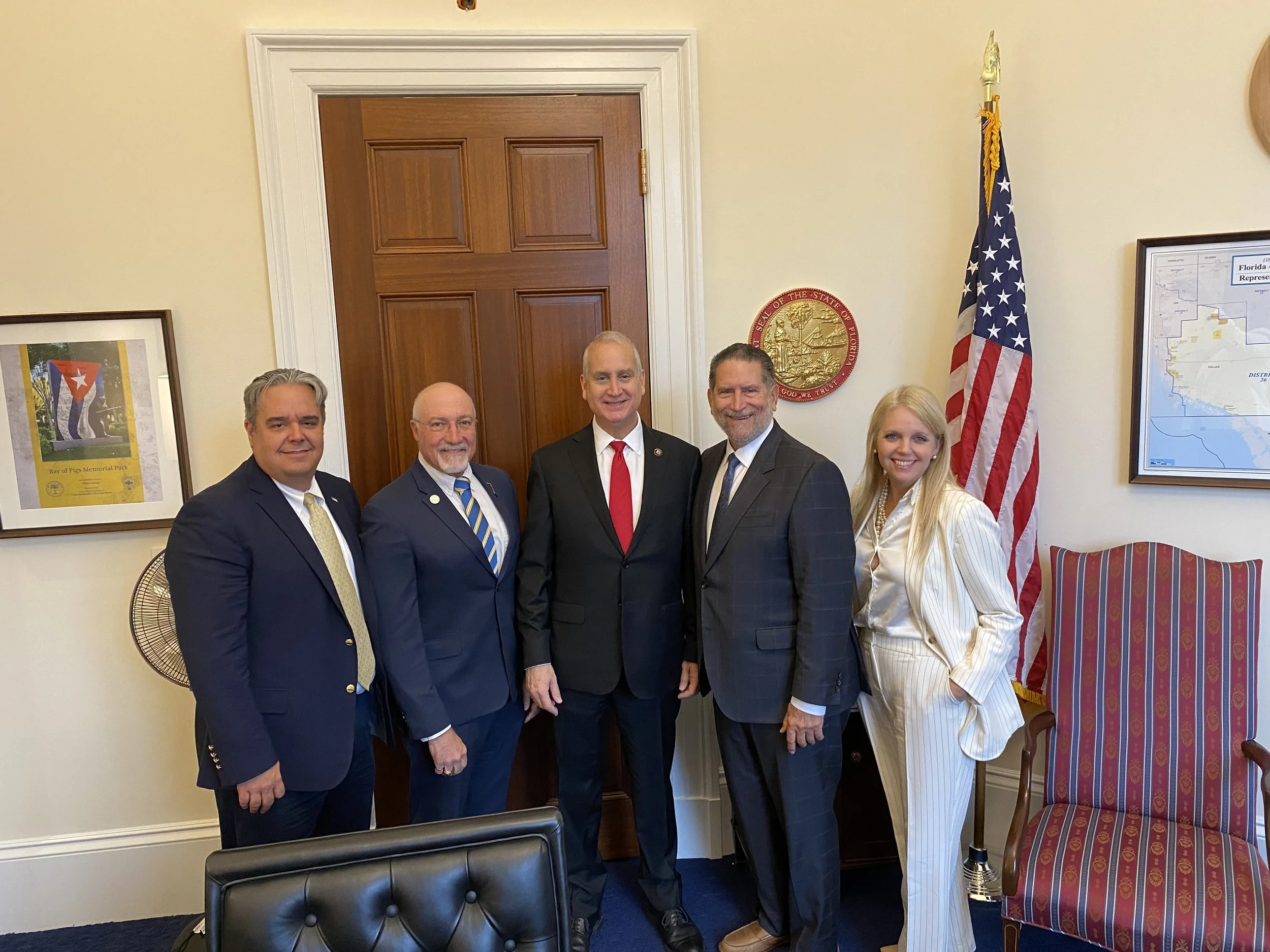 Group of five people standing inside a government office, with the U.S. flag and a state seal on the wall behind them, in a formal setting.