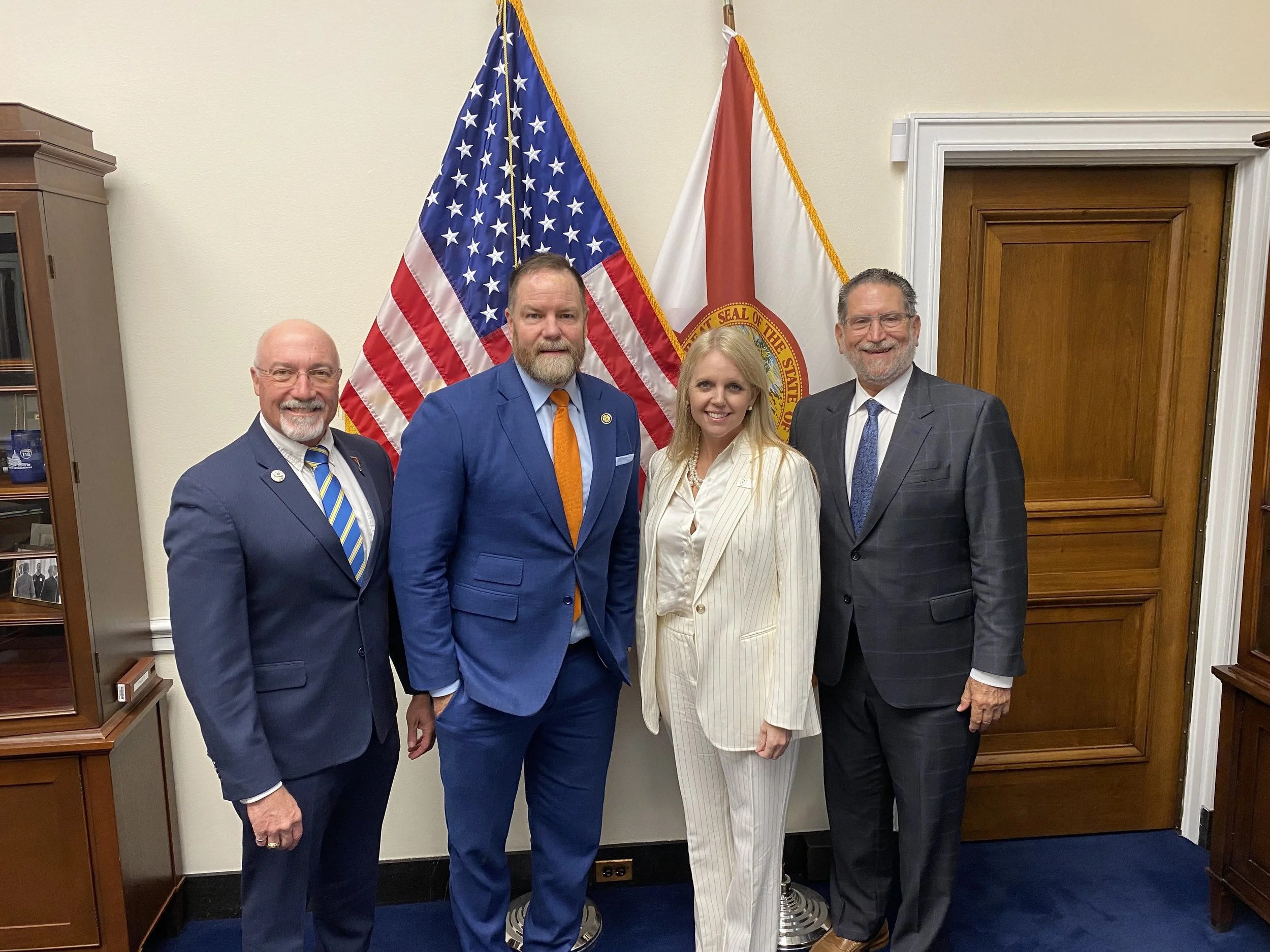 Four people standing together in front of American and Florida state flags, dressed in business attire, indoors.