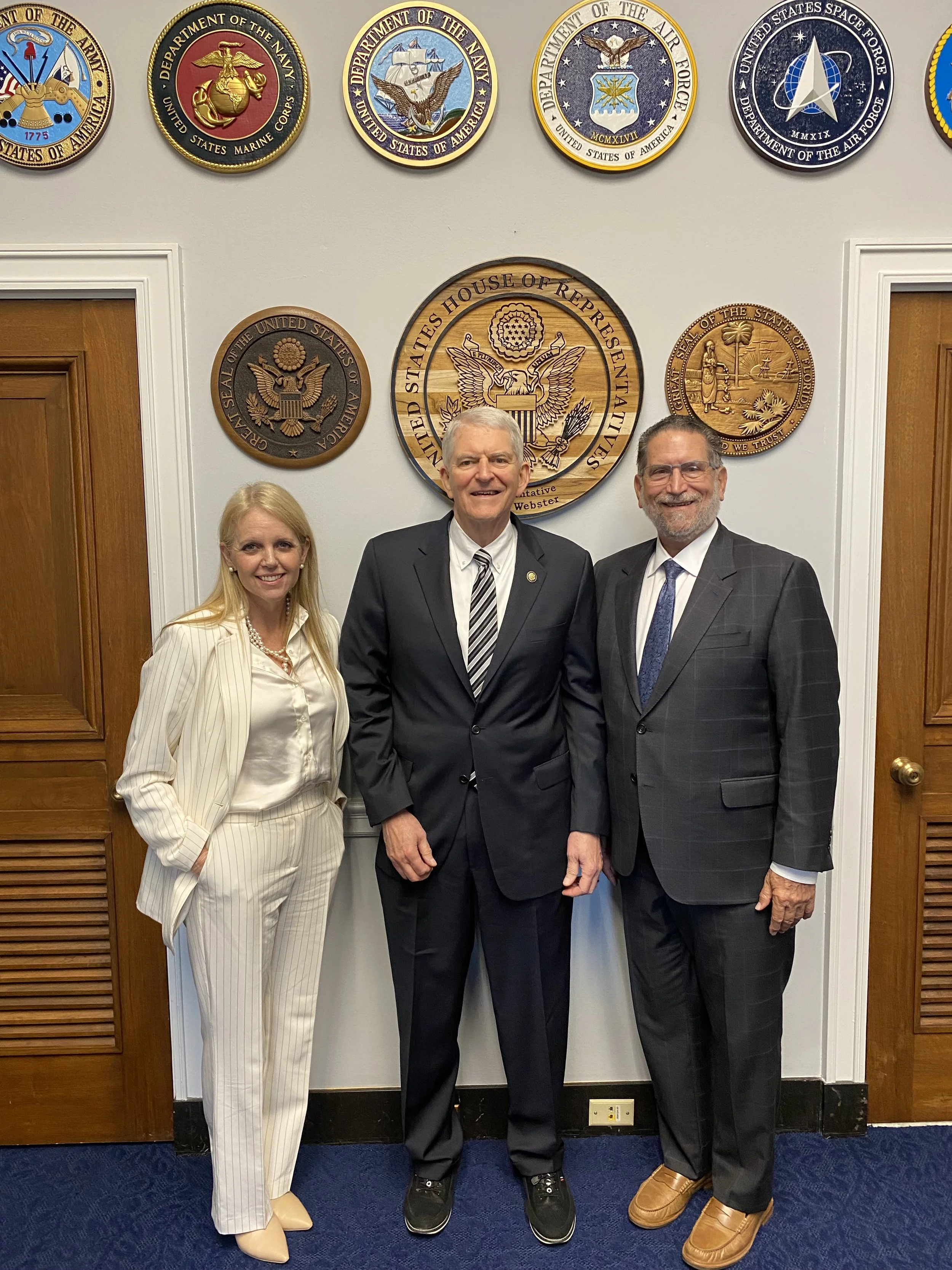 Three professionally dressed individuals standing in front of a wall decorated with seals of various United States government departments. The woman on the left is wearing a cream-colored pinstripe suit, the man in the middle is in a black suit with a striped tie, and the man on the right is in a checked gray suit with a dark blue tie. They are smiling and posing for the photo.