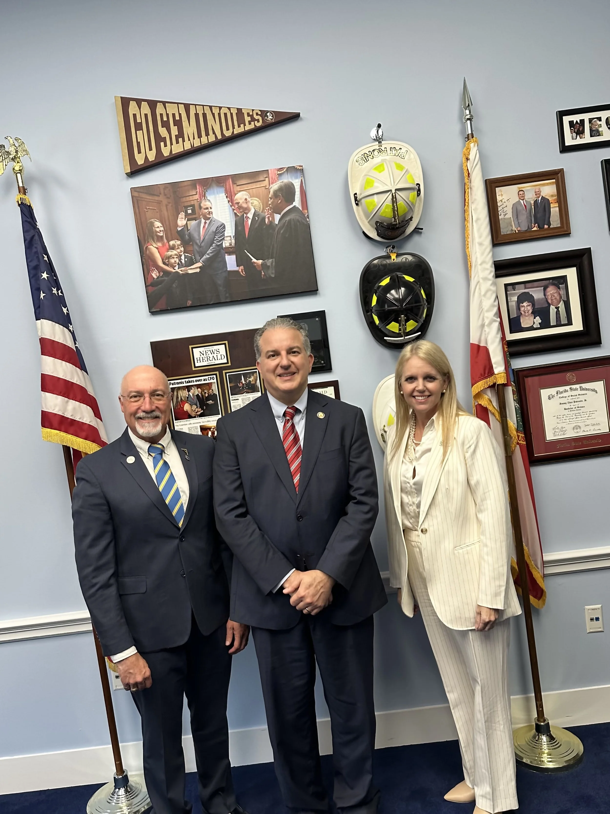 Three individuals in suits stand in front of a wall with flags, framed photos, and helmets, with a small crowd and a flag with 'GO SEMINOLES' banner in the background.