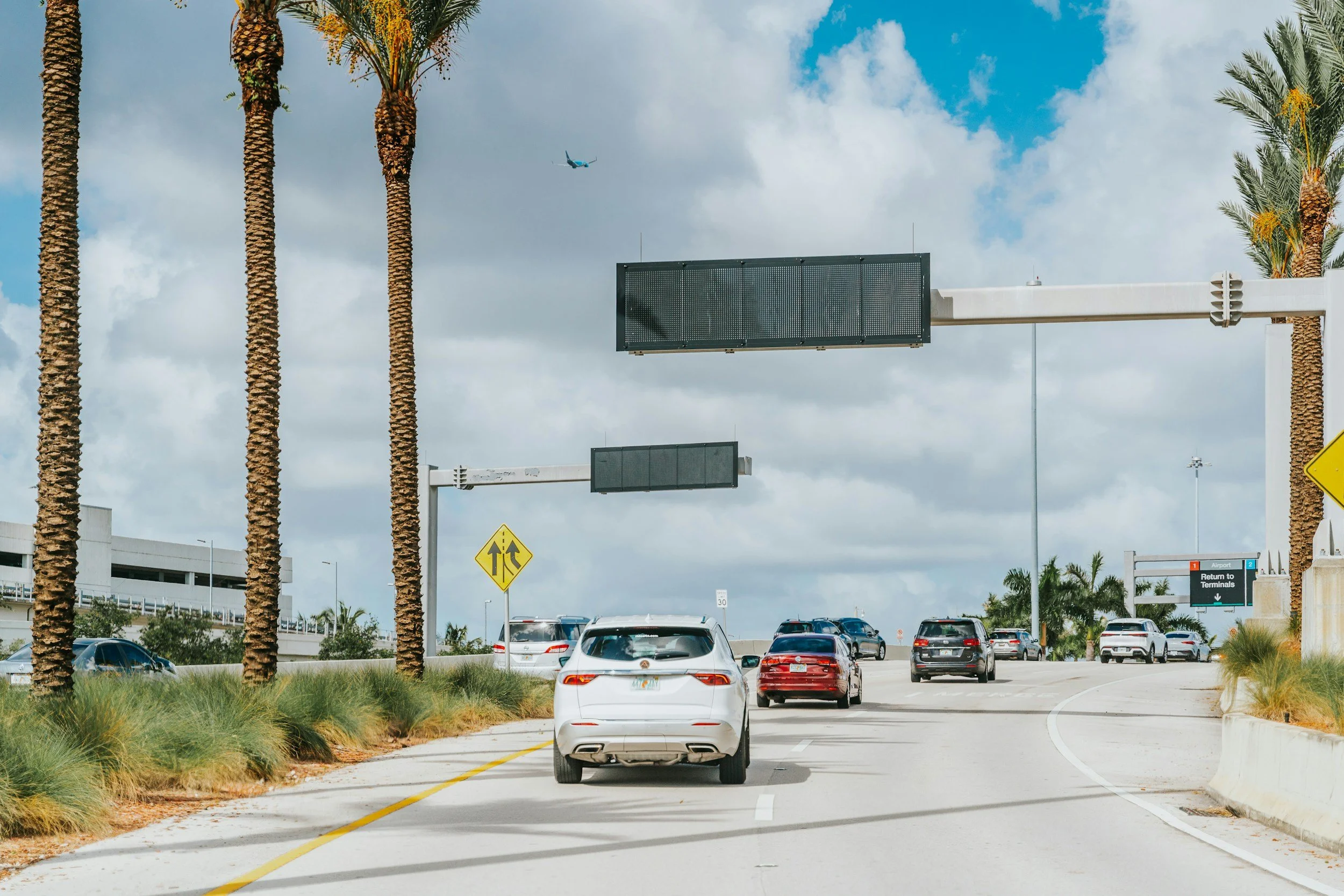 A road at an airport with multiple cars, palm trees lining the sides, yellow pedestrian crossing signs, and an overhead digital signboard against a partly cloudy sky, with an airplane flying in the background.