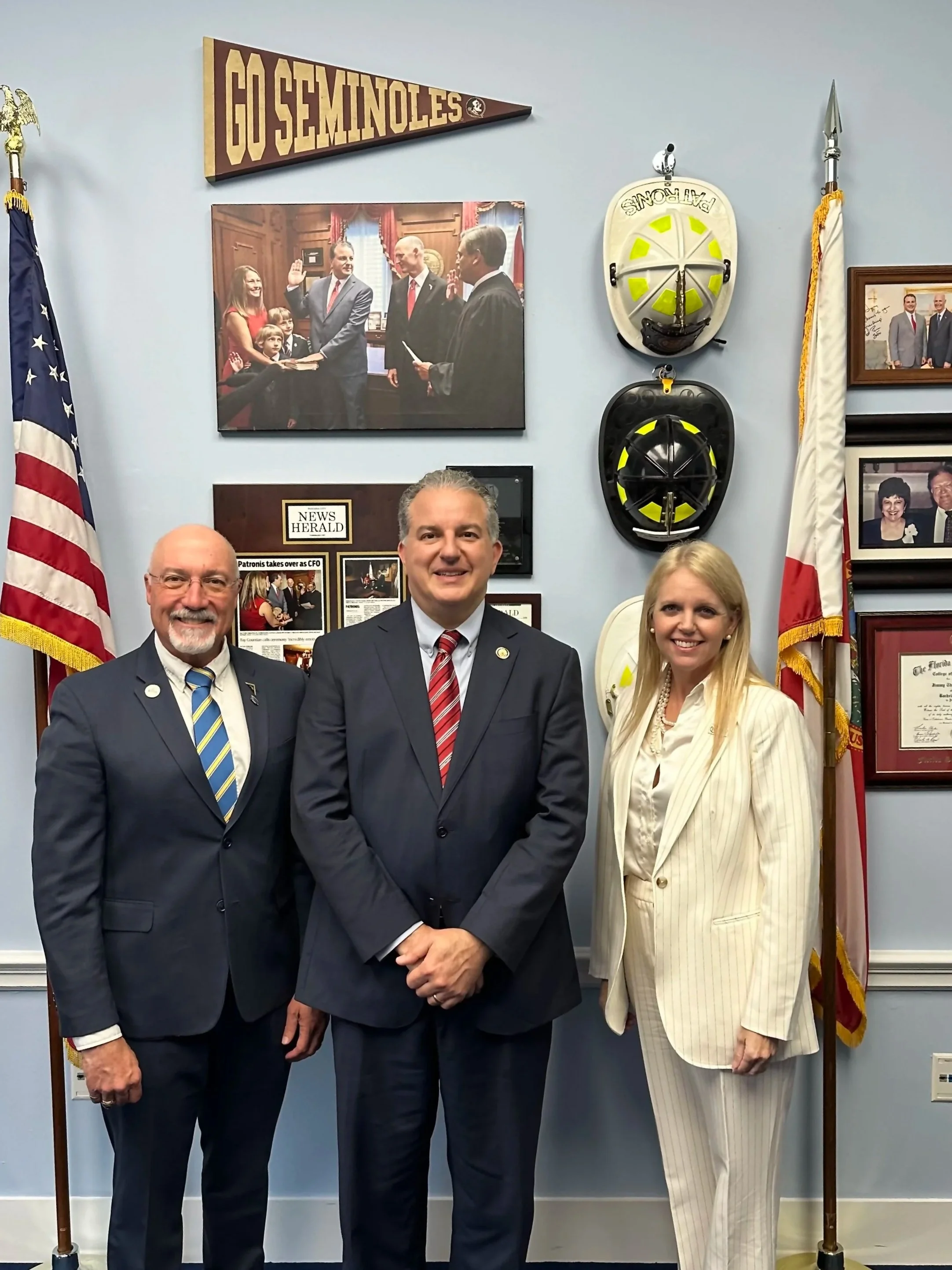 Three individuals in suits stand in front of a wall with flags, framed photos, and helmets, with a small crowd and a flag with 'GO SEMINOLES' banner in the background.