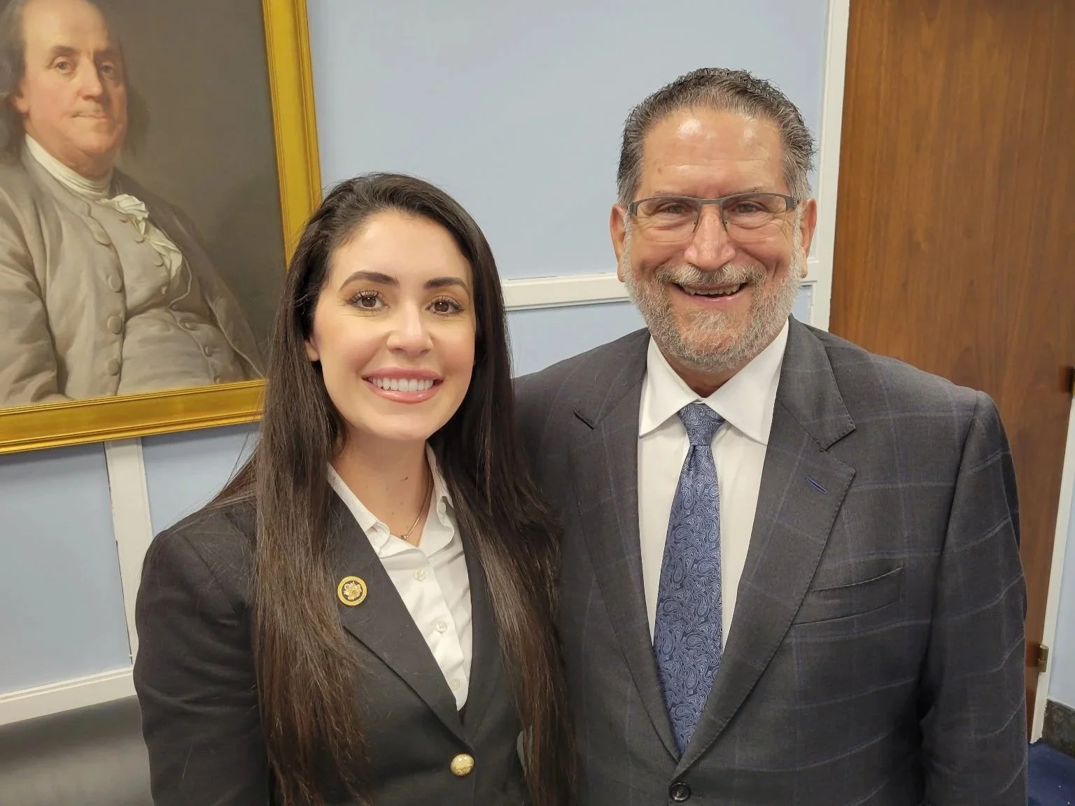 A young woman and an older man in suits are standing and smiling in a room with blue walls, a black leather couch, and a framed portrait of a man in historical clothing hanging on the wall behind them.