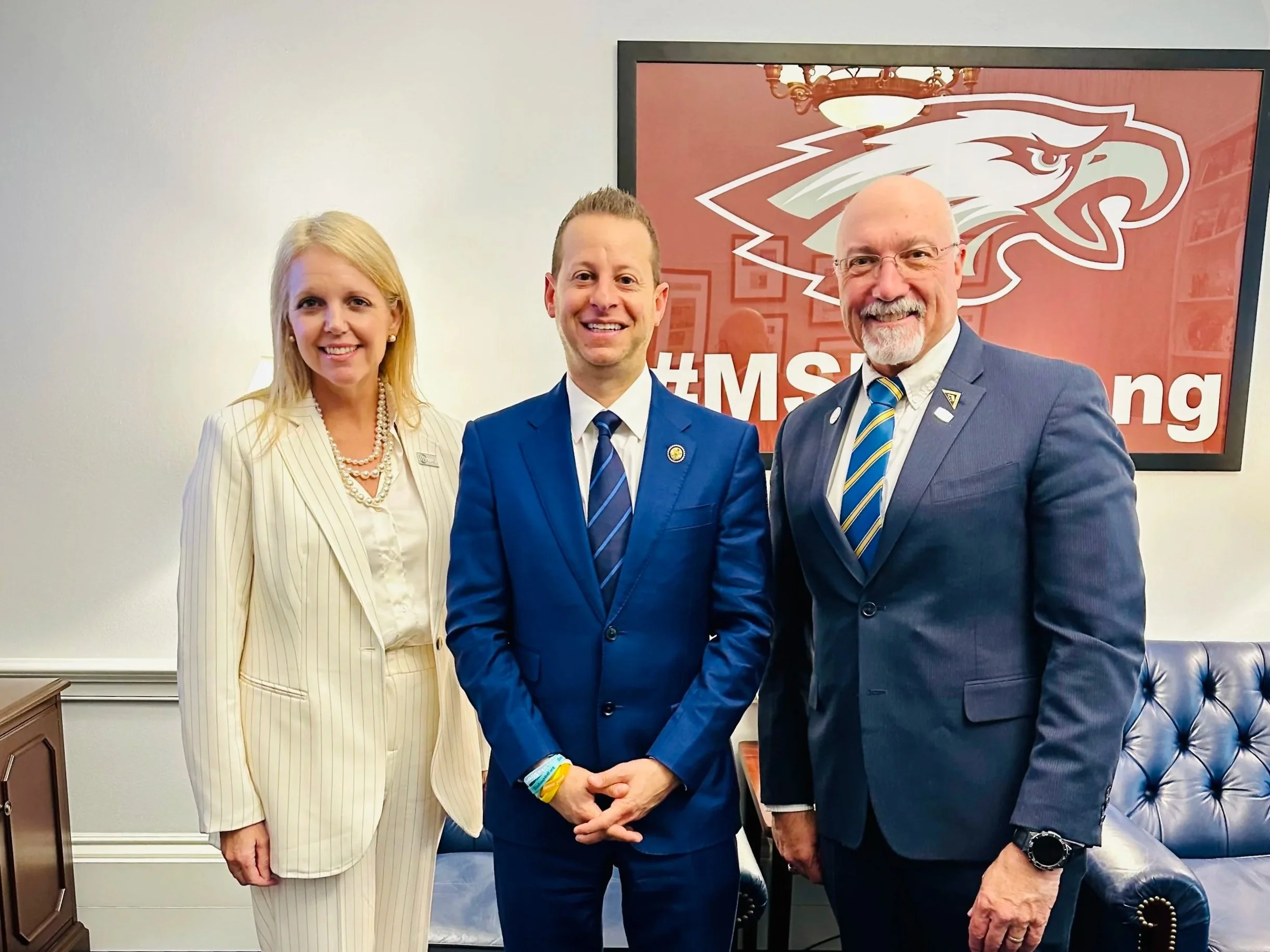 Three people in business suits standing in an office, smiling for a photo, with a large framed logo of a stylized eagle behind them and a hashtag #MSI in the background.