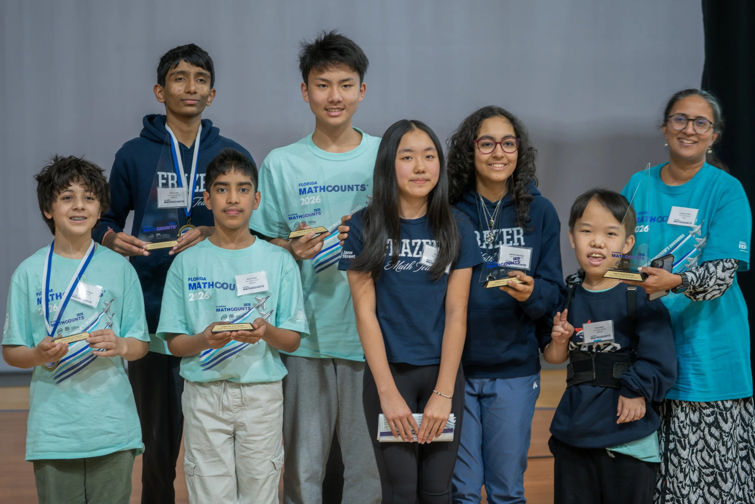 A group of children and a woman holding awards at a math competition, standing in front of a plain backdrop.