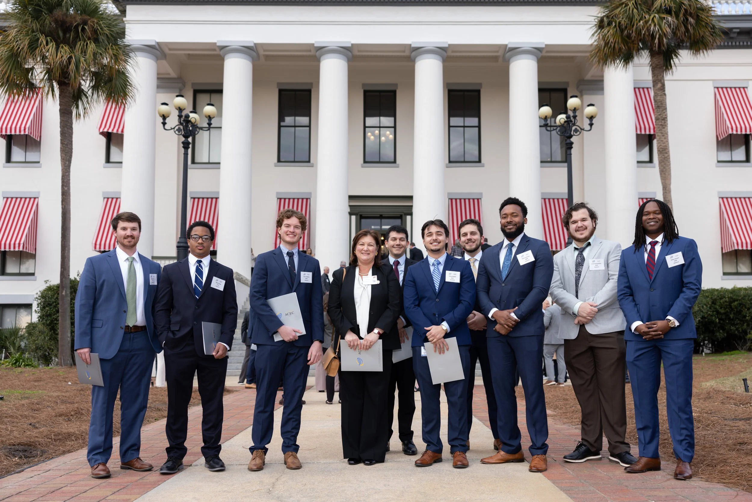 Group of nine diverse young professionals in formal business attire standing in front of a white building with red-striped awnings, palm trees, and lamp posts, smiling for a photo.