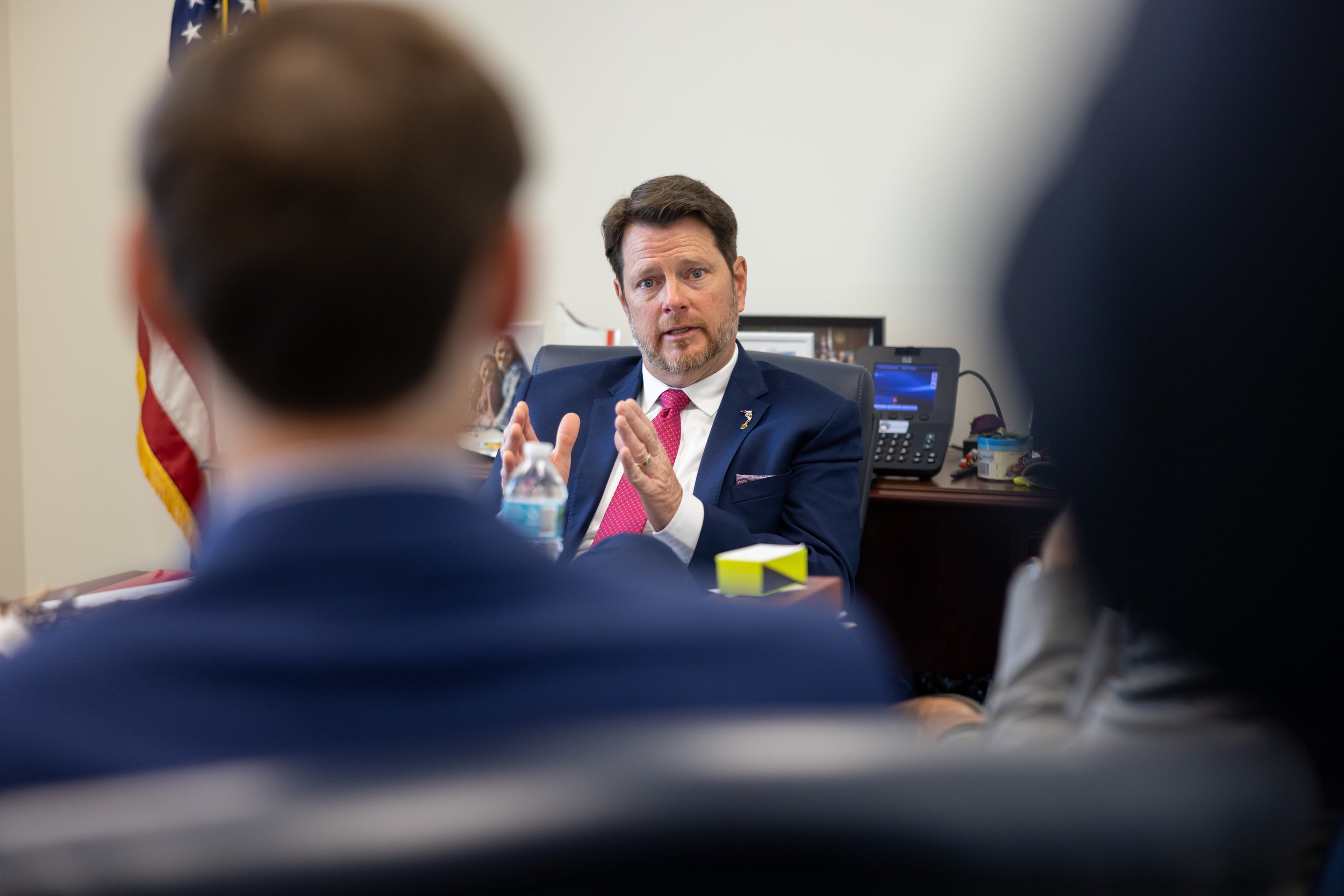 A man in a dark suit with a red tie is sitting at a desk, speaking and gesturing with his hands during a meeting. His back is to the camera, and the focus is on him.