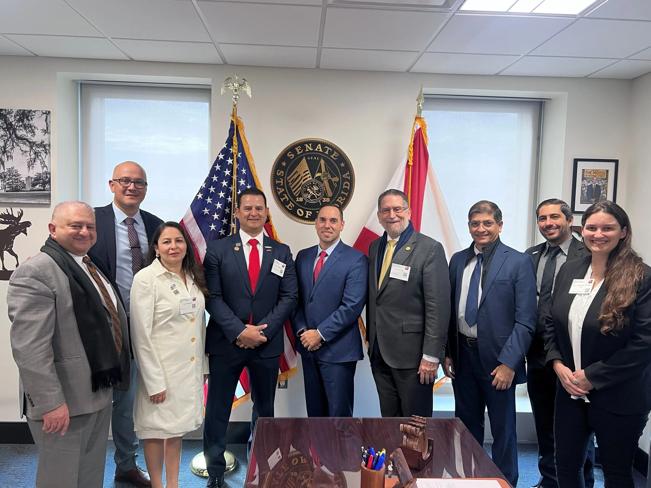 Group of ten people, including men and women, standing together in a government office with flags and a seal of the Florida Senate behind them.