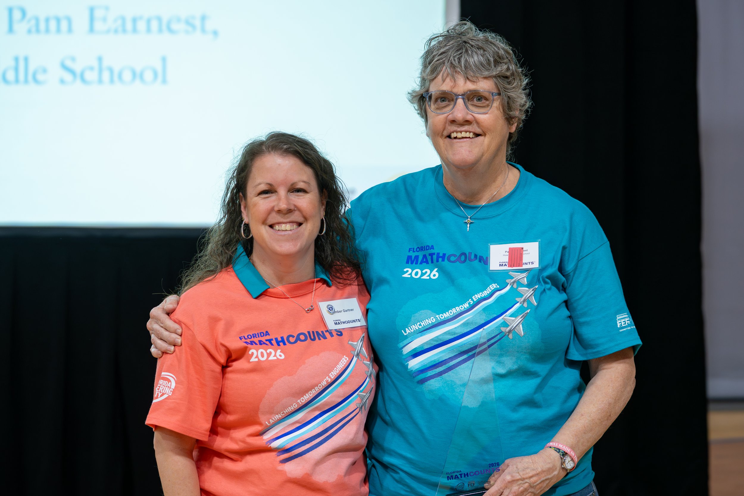 Two women standing together smiling, wearing Miami County 2026 T-shirts at a conference, with a presentation screen in the background.
