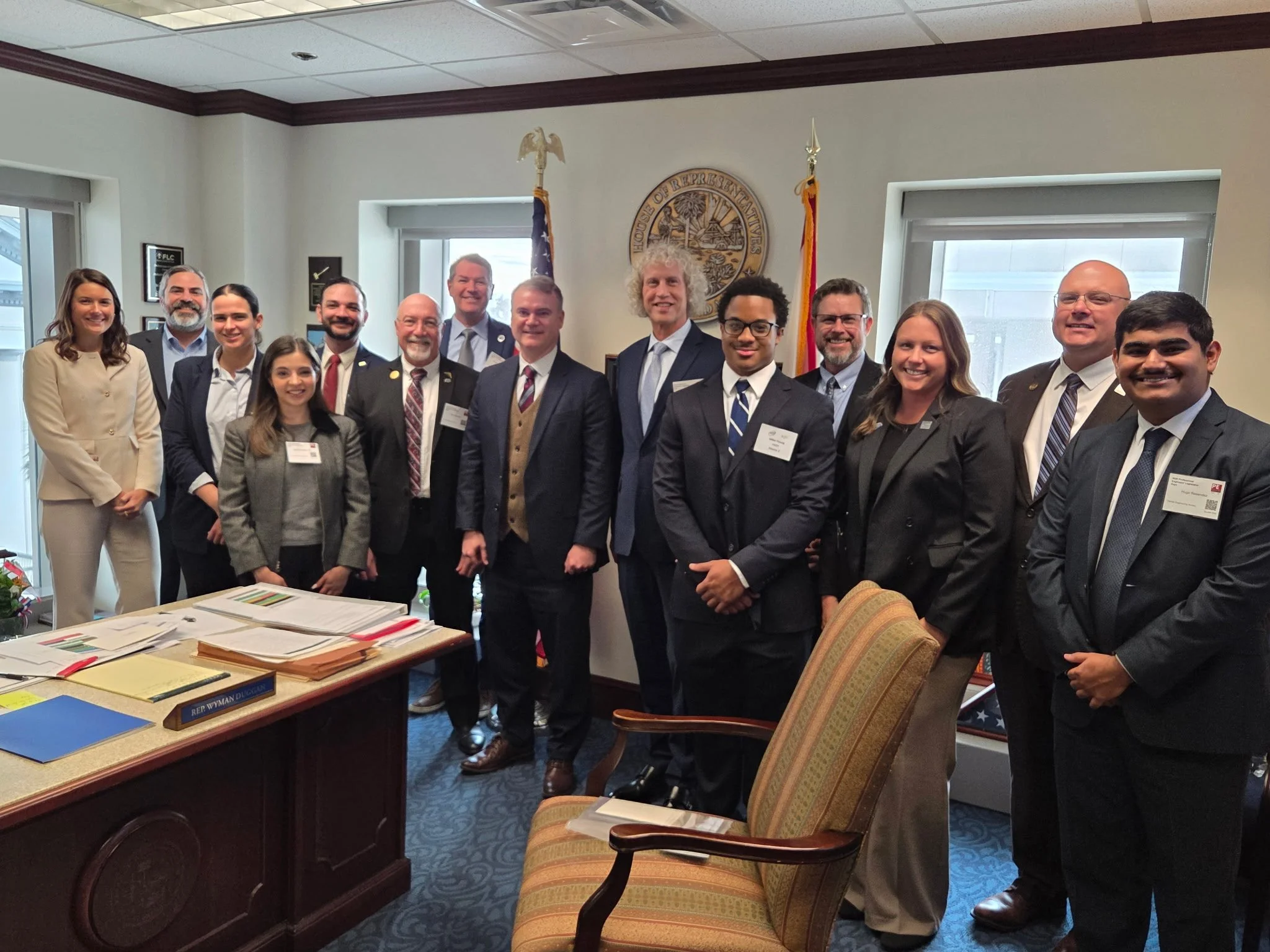 Group of 13 people in business attire posing for a photo in a room with a desk, flags, and a seal on the wall behind them.
