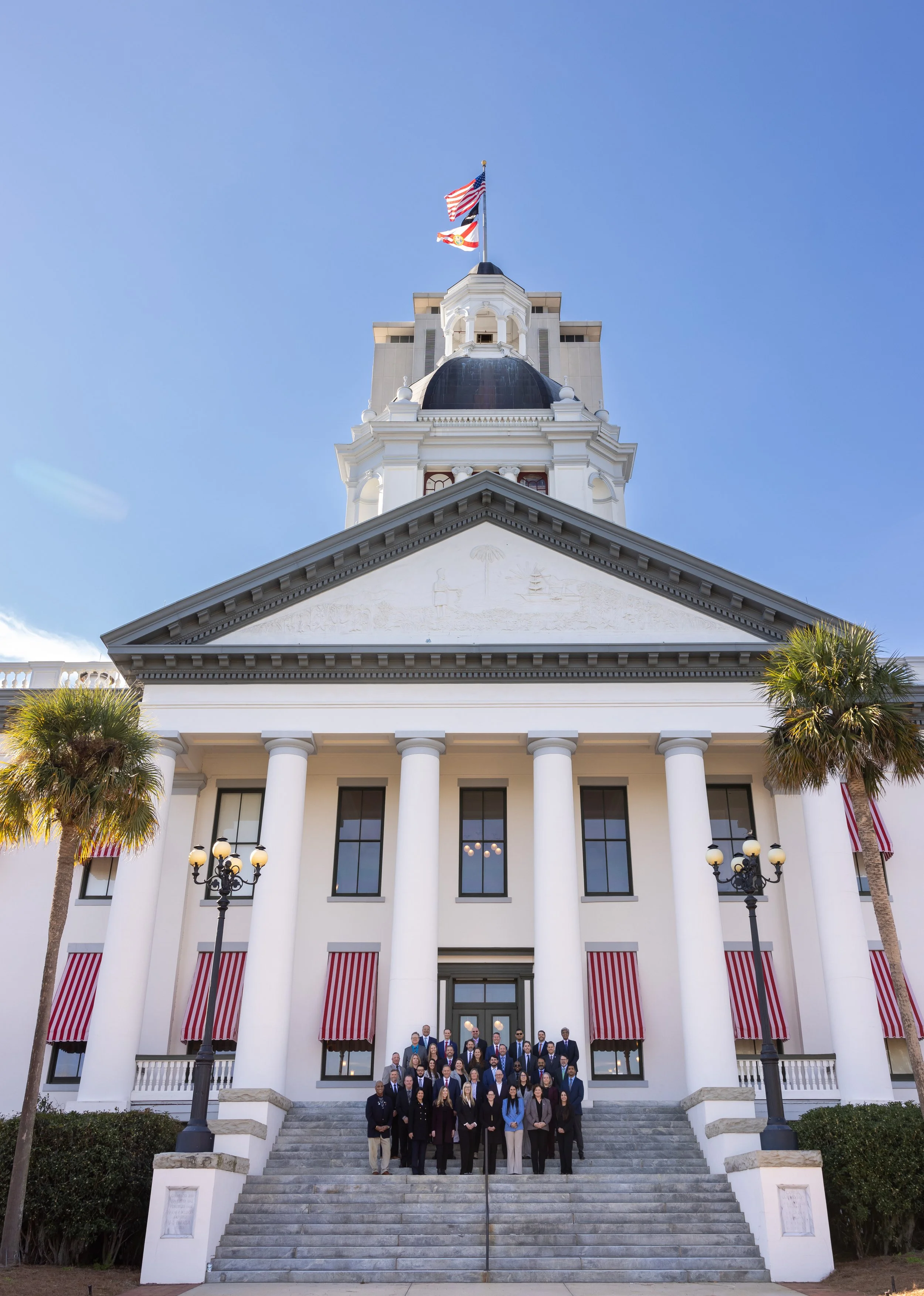 A group of people in formal attire standing on stairs in front of a large white government building with tall columns and striped red and white awnings, under a clear blue sky, with palm trees on either side.