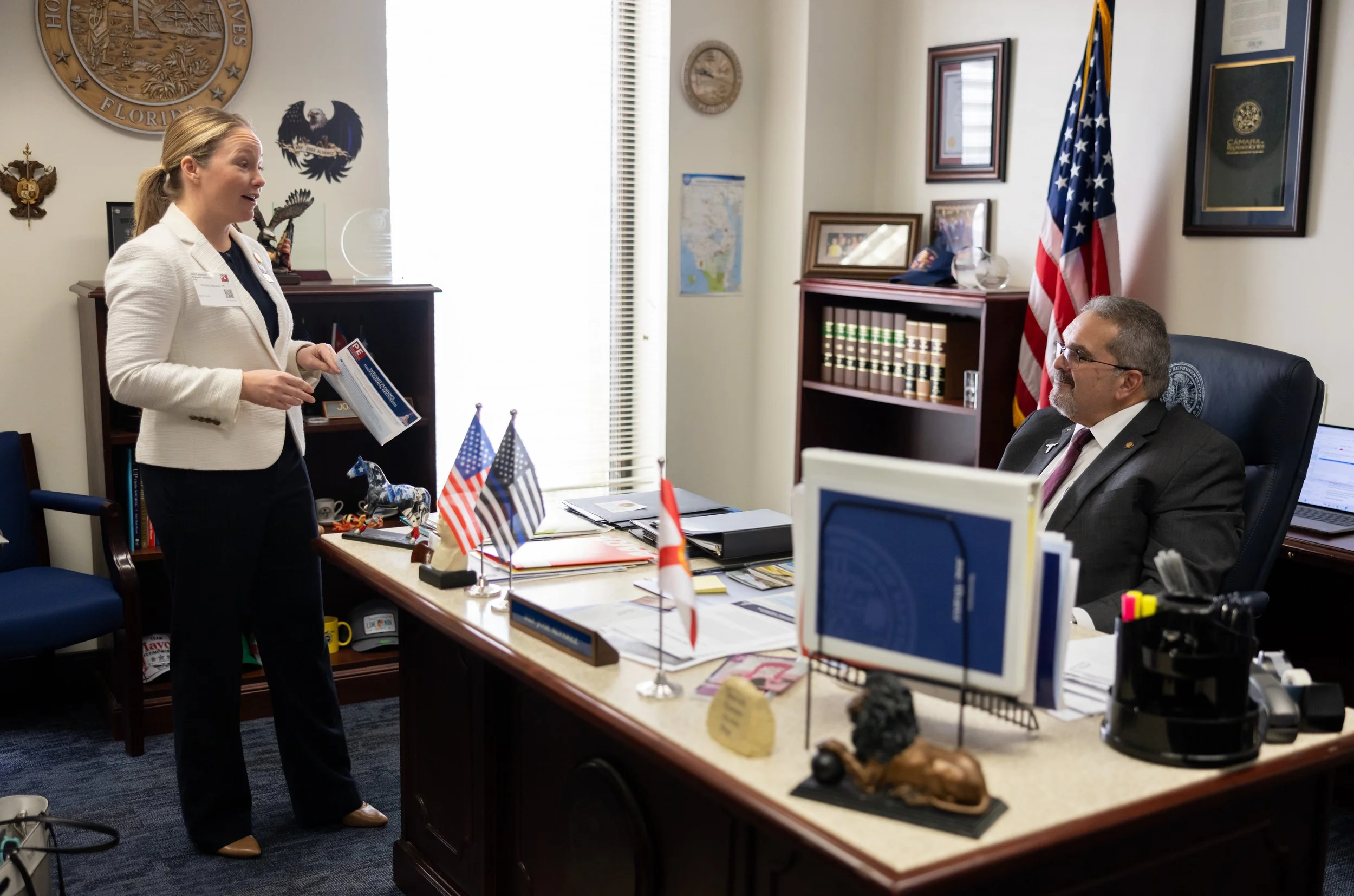 A woman standing and talking to a man sitting behind a desk in an office. The woman is holding papers and appears to be explaining. The office has flags, books, and framed pictures on the walls.