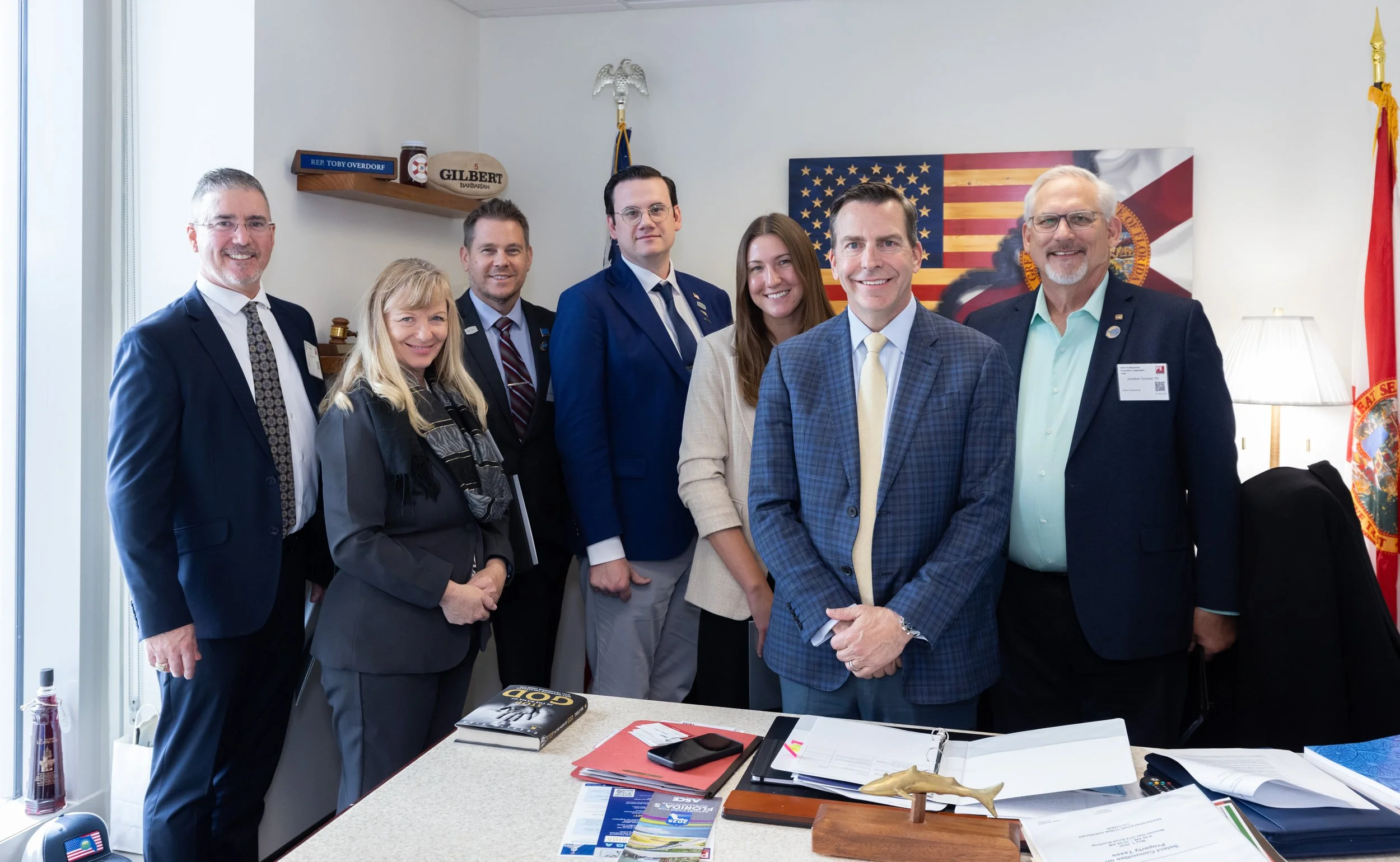 A group of seven people dressed in business attire posing in an office, with American and Florida flags in the background. They are smiling and standing around a desk with documents, a magazine, and a small decorative shark.