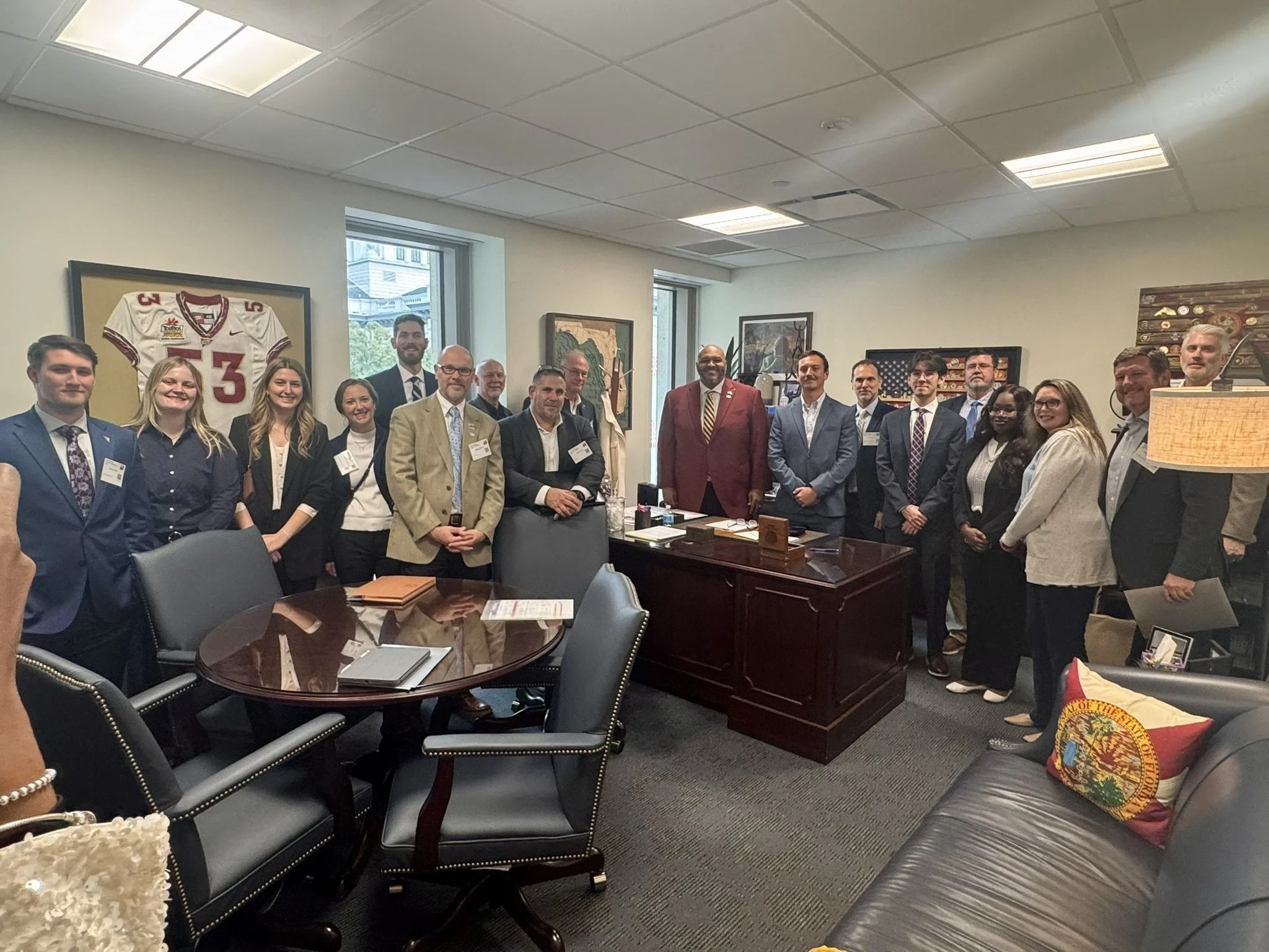 Group of professionals in business attire posing in a conference room, with framed artwork and a football jersey on the wall.