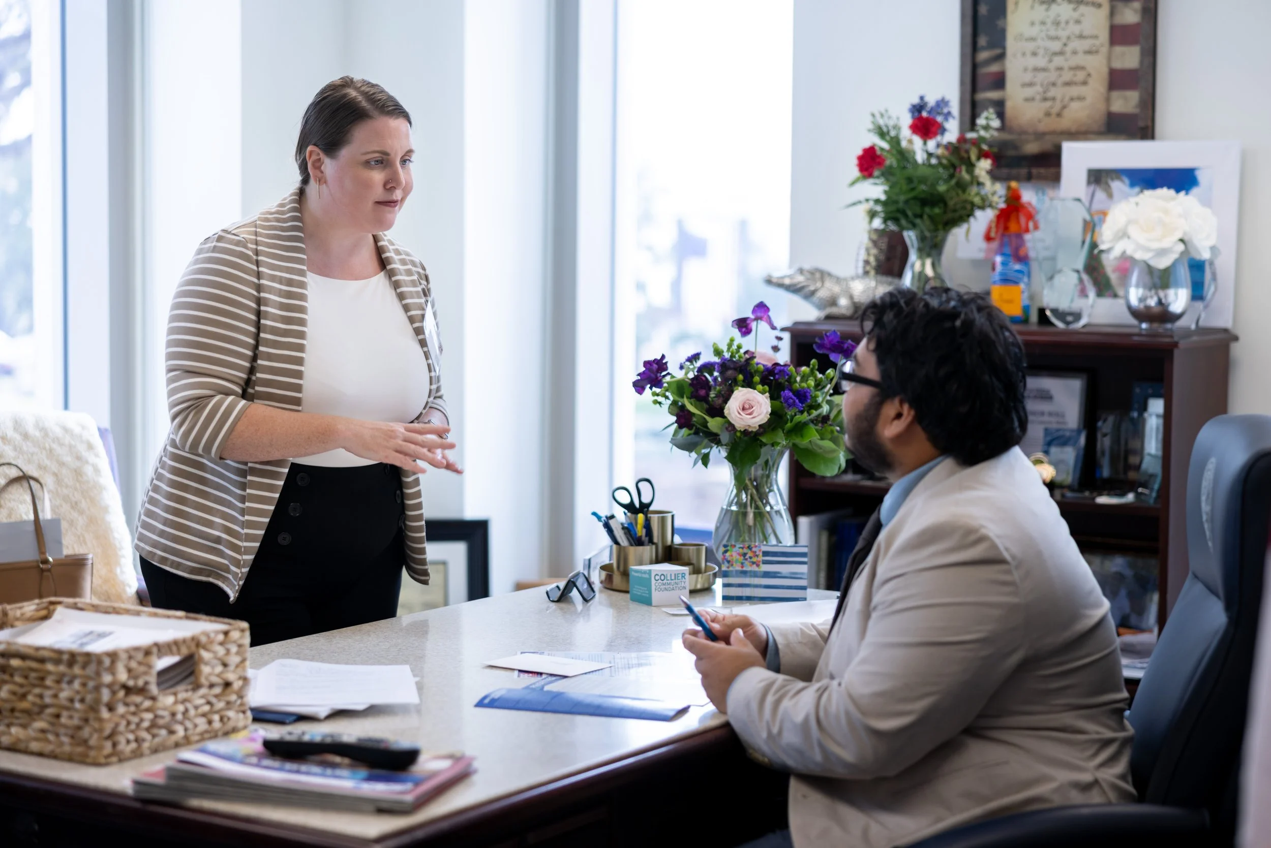 A woman standing and talking to a man sitting at a desk in an office. The desk has papers, a pen, and a colorful vase with flowers. The office has bookshelves, framed photos, flowers, and decorative items.