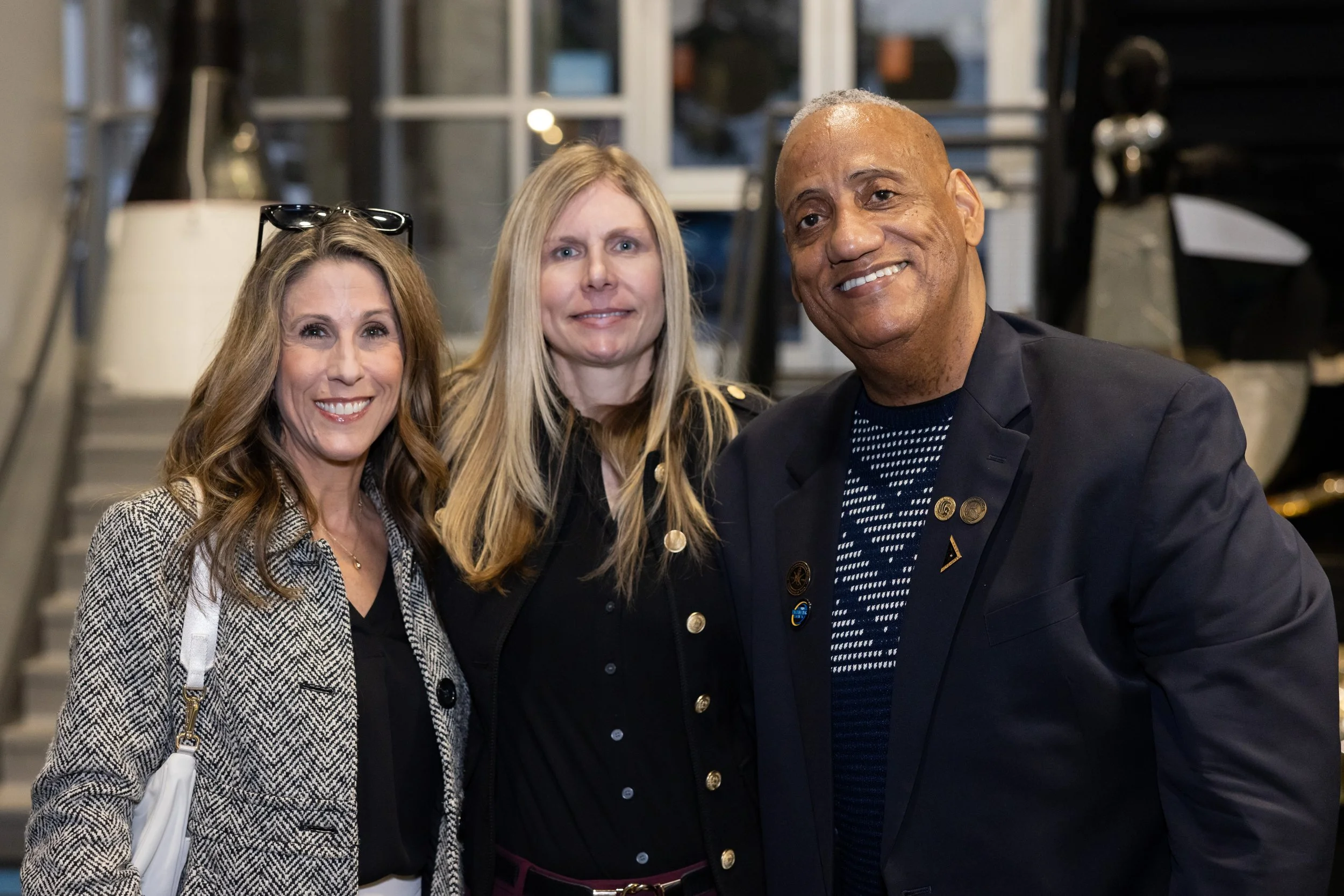 Three people standing together, smiling, in a well-lit indoor setting with stairs and display items behind them.