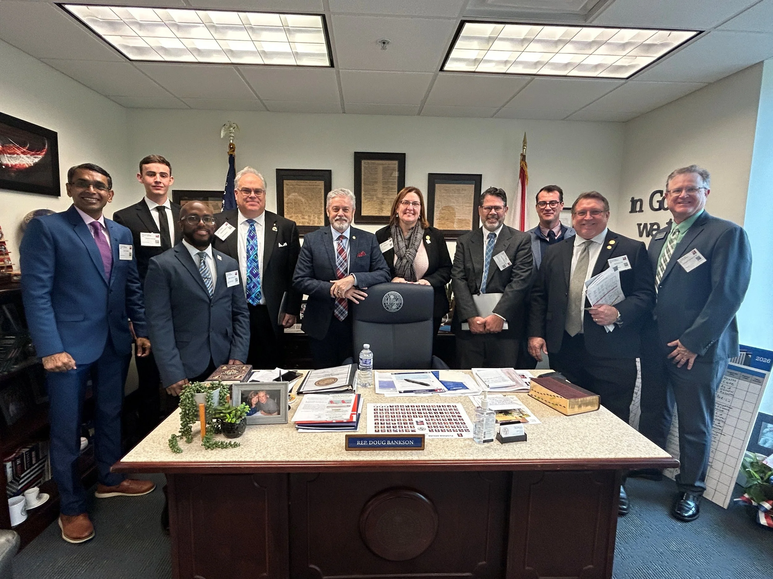 Group of eleven people in business attire gathered in an office around a desk, with framed documents on the wall behind them. The desk has various papers, a water bottle, and a framed photo. A nameplate on the desk reads 'Rep. Doug Bankson'.