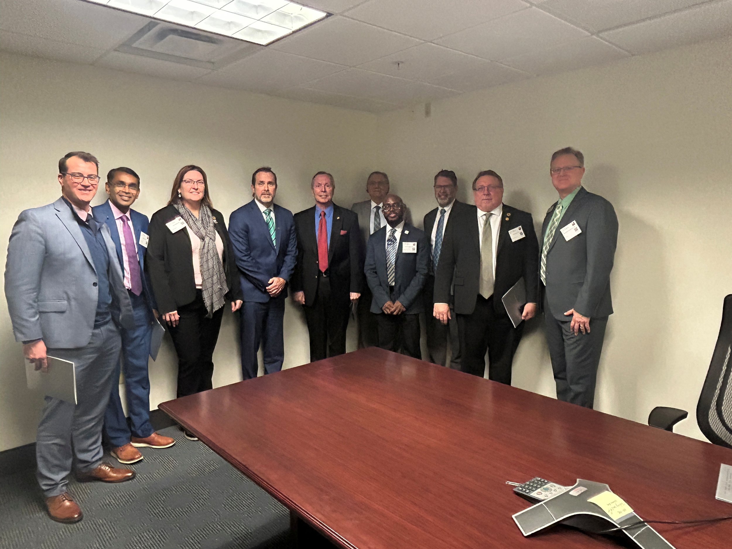 A group of eleven professionally dressed individuals standing in a conference room, posing for a photo. The setting is an office with a large wooden table and a telephone in the foreground.