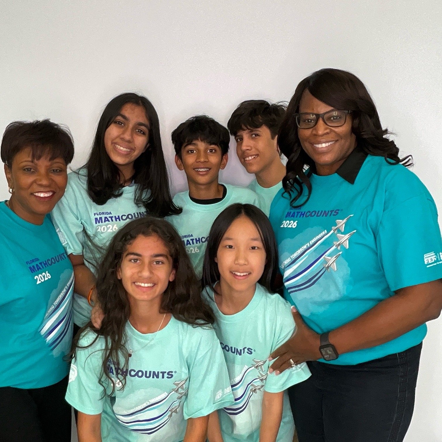 Group of seven people, including children and adults, wearing matching teal and turquoise T-shirts with 'MATHCOUNTS 2026' printed on them, standing in front of a plain white wall and smiling at the camera.
