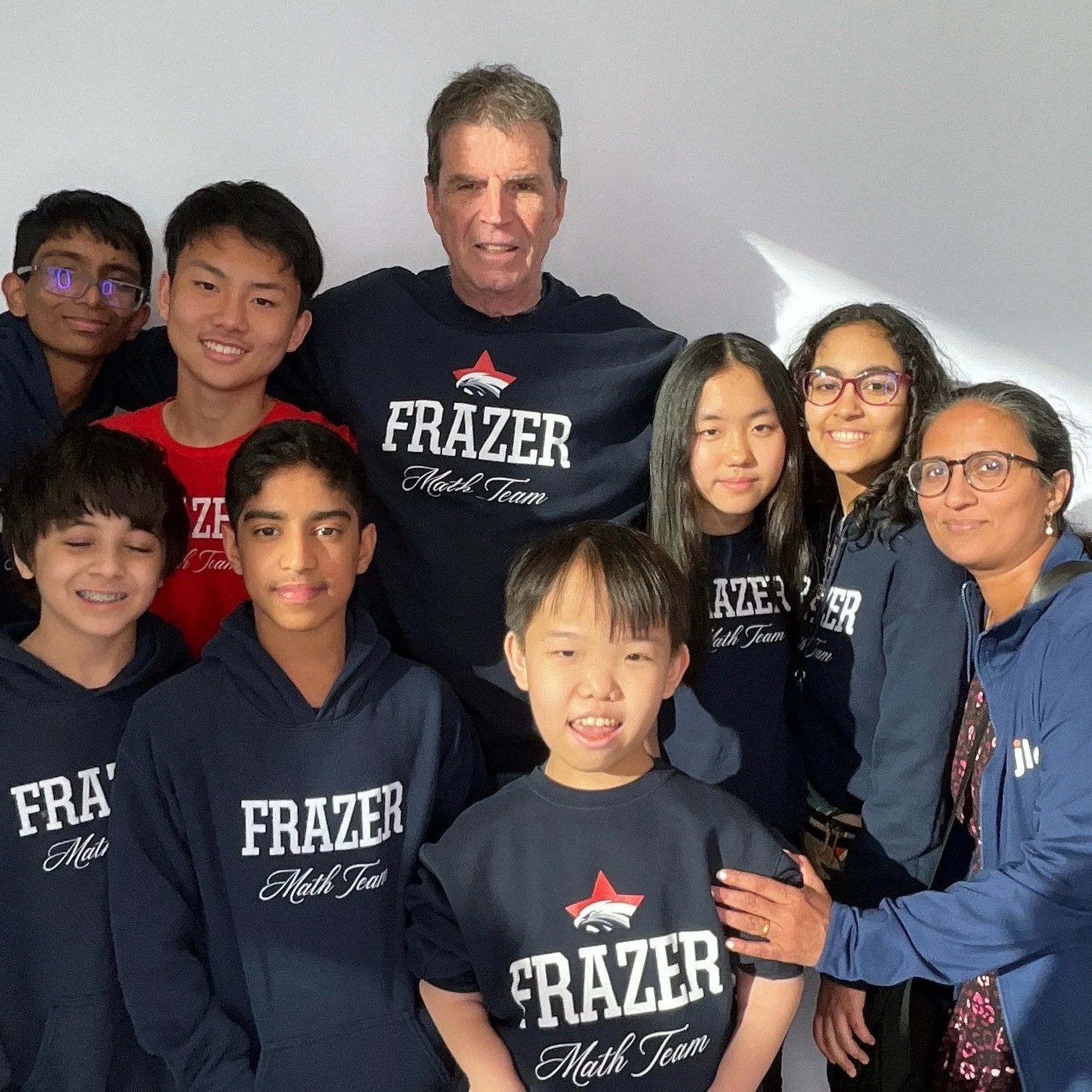 Group of young students and two adults posing together, all wearing navy blue or red hoodies with 'FRAZER Math Team' written on them, standing indoors against a plain white wall.