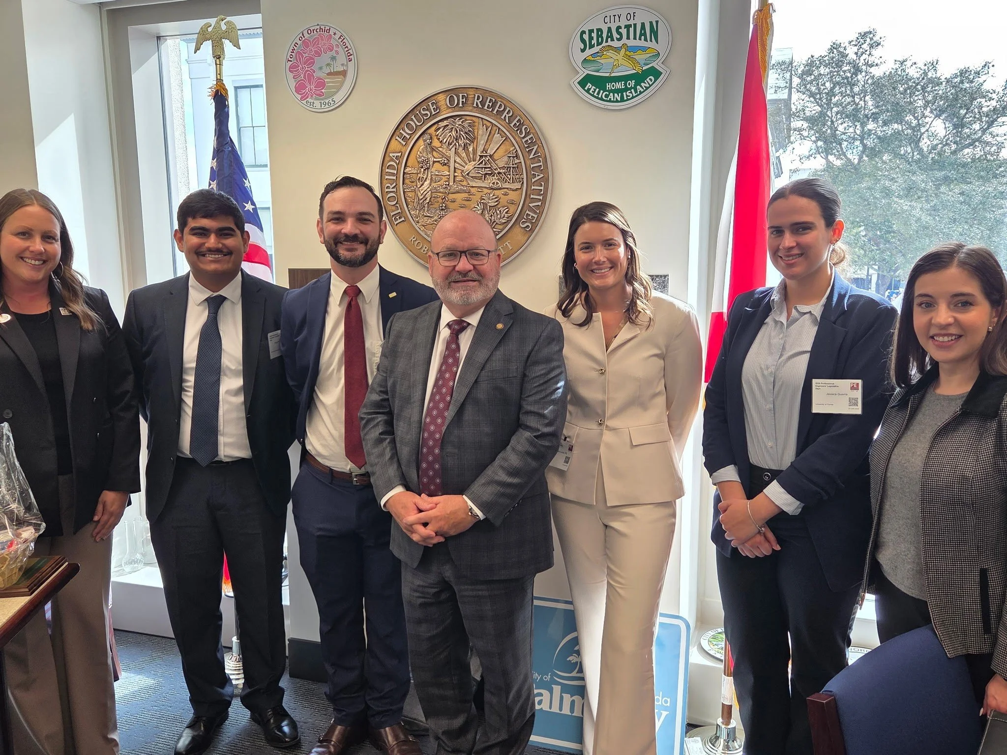 Group of seven people standing in front of a wall with government seals and flags, smiling for a photo.
