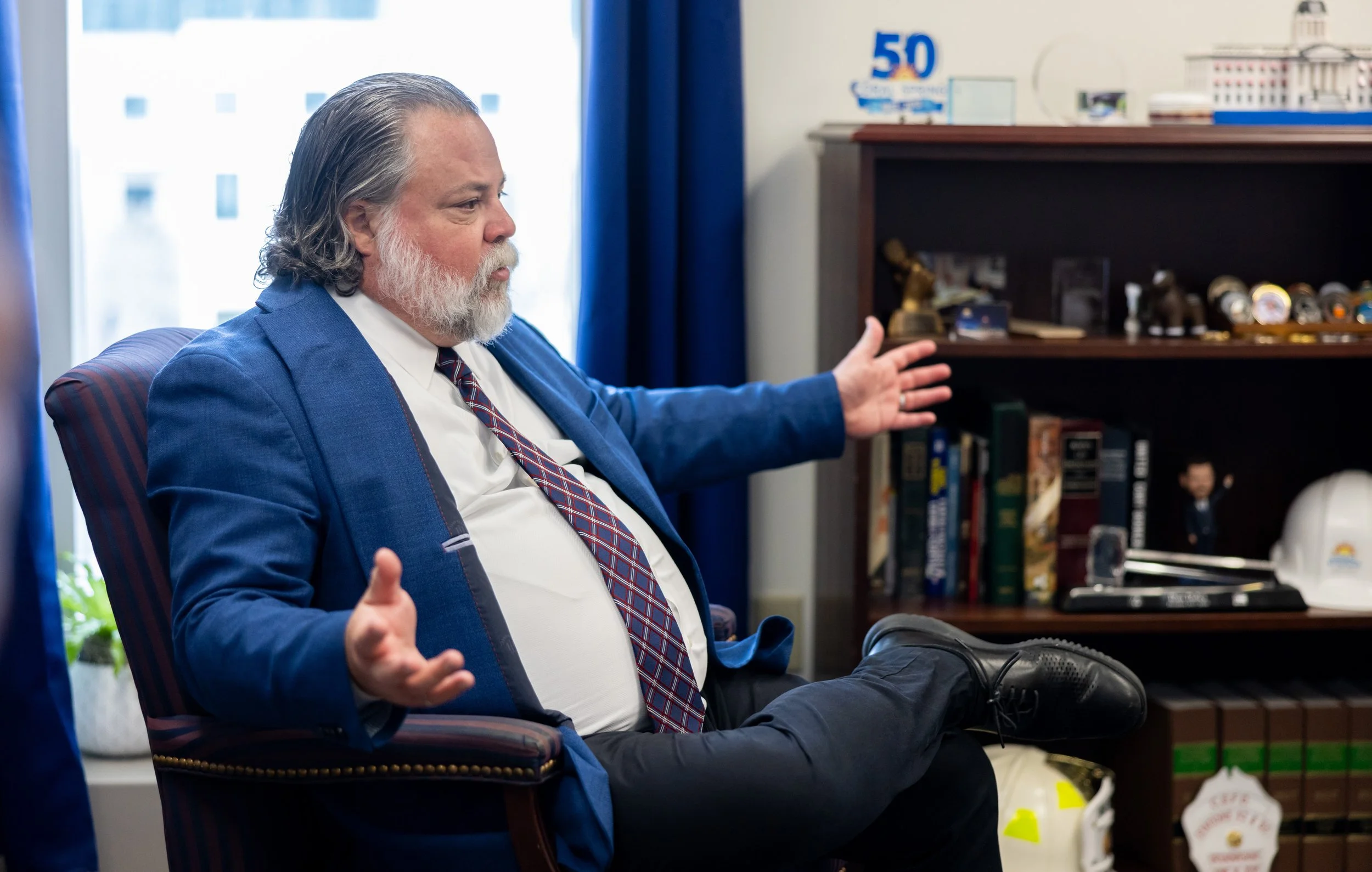 A man with gray hair and a beard, dressed in a blue suit, white shirt, and patterned tie, sits in a chair with his legs crossed, gesturing with one hand, in an office with shelves filled with books, decorative items, and a '50' anniversary sign.