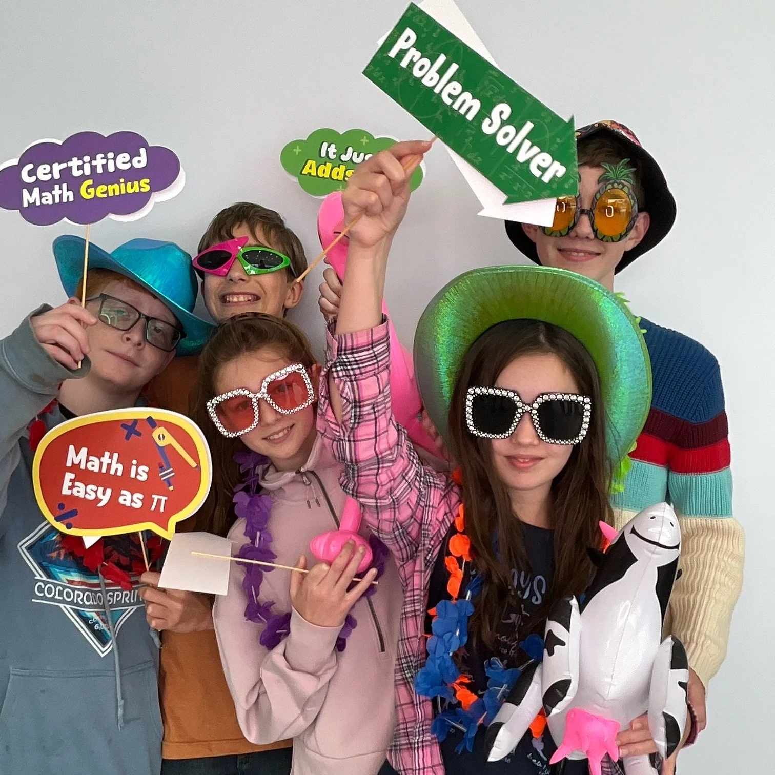 Group of children celebrating with colorful hats, sunglasses, and props such as signs and inflatable animals, wearing leis, and holding various signs including one that says 'Problem Solver' and another that says 'Math is Easy as Pi', against a plain