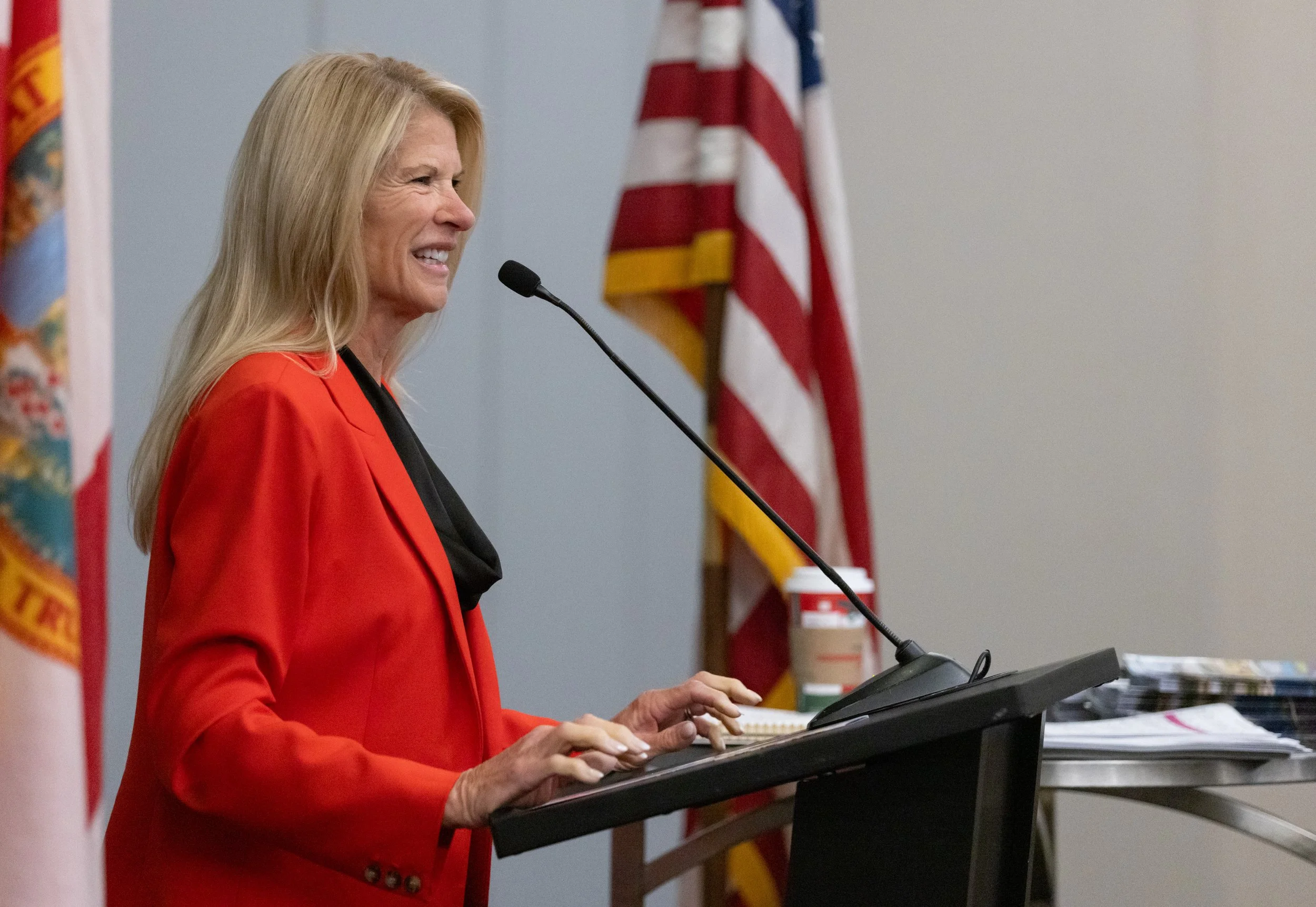 A woman with blonde hair in a red blazer speaking at a podium with a microphone, with an American flag and another flag in the background.