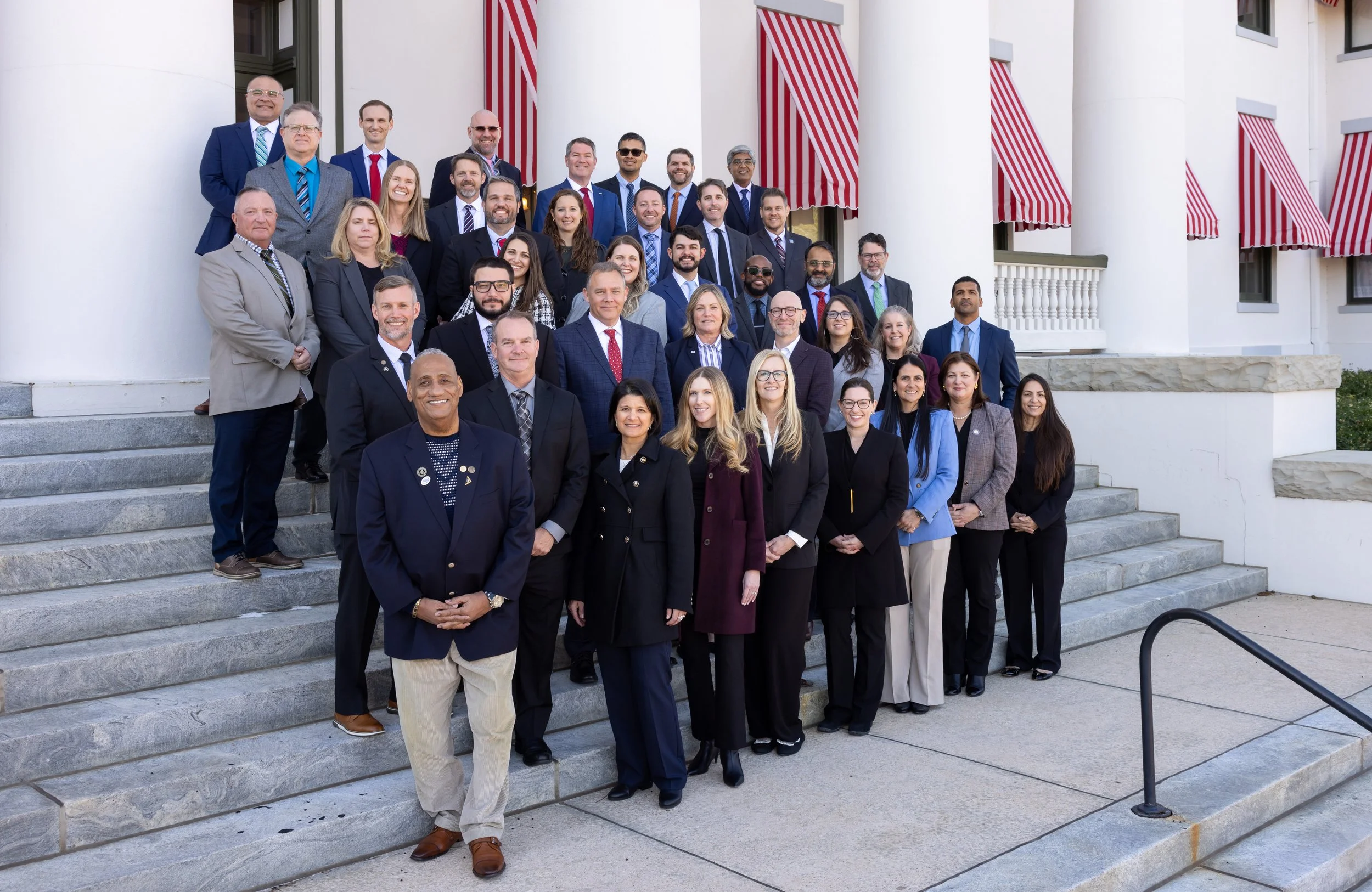 Group of professionally dressed people standing on steps outside a white building with red-striped awnings, posing for a photo.