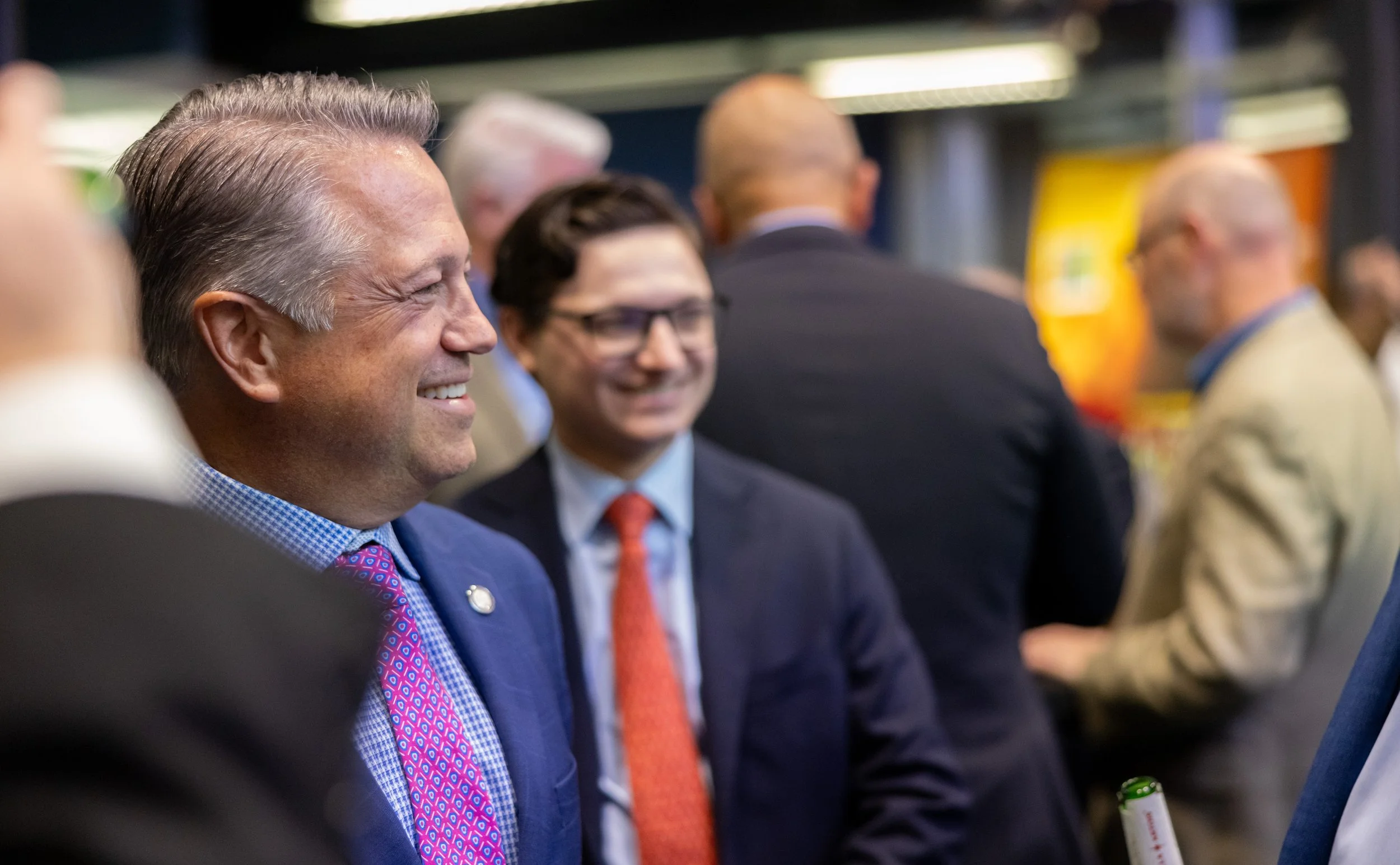 Group of men in suits and business attire at a professional event, smiling and engaging in conversation.
