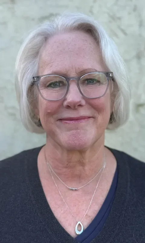 A smiling older woman with short gray hair and glasses, wearing layered necklaces and a dark top, standing outdoors against a light-colored stone wall.